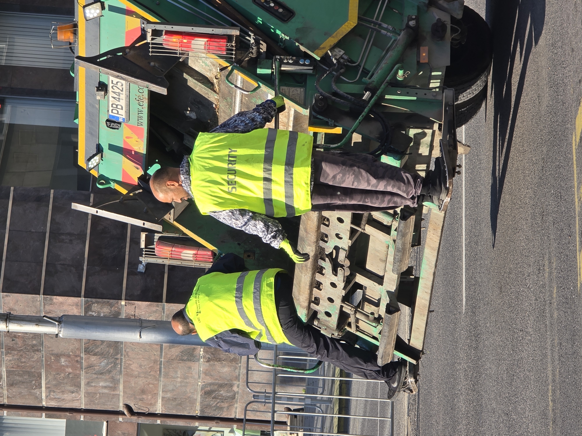 Two male sanitation workers are standing on the back of a green garbage truck. The worker on the left is wearing black pants, a blue shirt, and a bright yellow vest with reflective stripes. He is facing away from the camera, with his back to the left. The worker on the right is wearing camouflage pants and a bright yellow vest with "SECURITY" printed on the back in black letters, along with reflective stripes. He is also wearing a patterned shirt underneath. He is facing away from the camera, looking towards the right. Both workers have dark hair and appear to be in their 30s or 40s. The garbage truck has a license plate that reads "PB 4425" and the website "www.efe.co" is visible on the side. The truck is parked on a paved road with yellow and white lines. In the background, there is a modern building with a dark tiled facade and a metal fence. The scene appears to be a street in Plovdiv, Bulgaria.