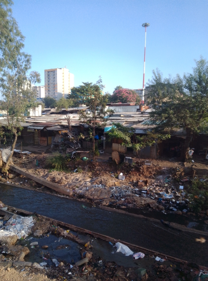The image displays an informal settlement situated in Nairobi, Kenya, specifically resembling an area along the Mathare River.
The foreground features a narrow, dark-colored river or canal flowing from left to right, heavily contaminated with solid waste, including numerous plastic bags, bottles, and miscellaneous debris accumulated on both banks and within the water. A diagonal concrete beam or support structure extends from the left bank into the river. The banks themselves are uneven and composed of dirt and mud.
The midground consists of a dense cluster of makeshift structures, predominantly constructed from corrugated metal sheets that exhibit significant rust and wear, alongside various other materials such as wood. These structures feature low-pitched, corrugated roofs. Piles of refuse and discarded materials are scattered throughout the ground between the buildings. Sparse vegetation, including small trees and shrubs, grows amidst the structures and along the banks. A single individual, wearing a blue shirt, is partially visible within the entrance of one of the structures located on the right side of the midground.
The background presents a clear blue sky. On the left, a multi-story, light-beige high-rise building is visible above the settlement, along with parts of other urban buildings. To the right of center, a tall, slender mast, painted with alternating red and white horizontal stripes, rises prominently, topped by a cluster of lights or antennae. Deciduous trees, some displaying pink blossoms, are also visible along the horizon line. The scene is illuminated by natural light, indicating either an early morning or late afternoon sun angle.