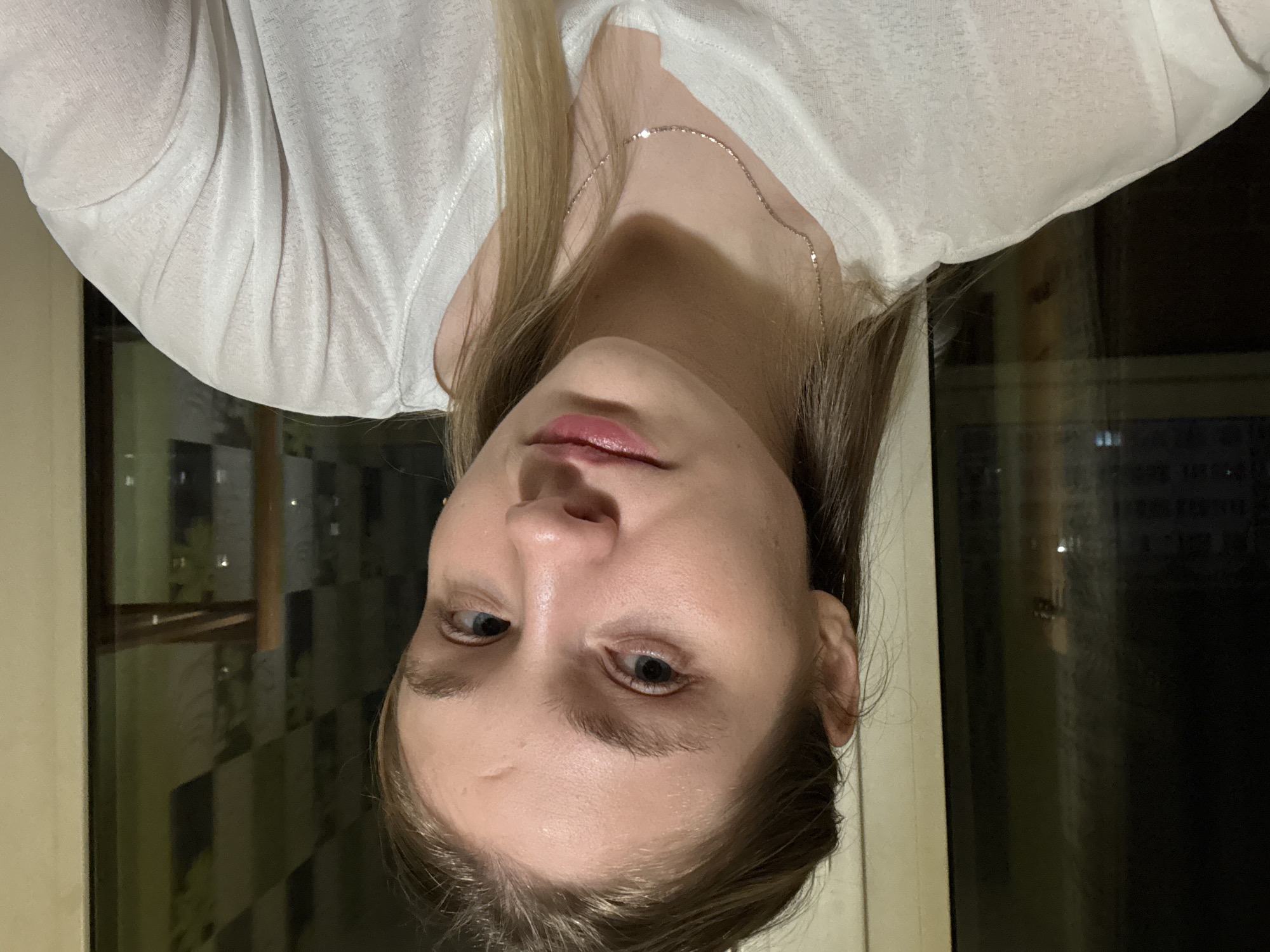 A woman with blonde hair and a silver necklace wears a white shirt and looks at the camera. She is indoors near a window, and lights are visible outside, possibly indicating a night scene. Based on the reflections in the window, it seems to be a residential area. There is no indication of the location being in Izhevsk, Russia.