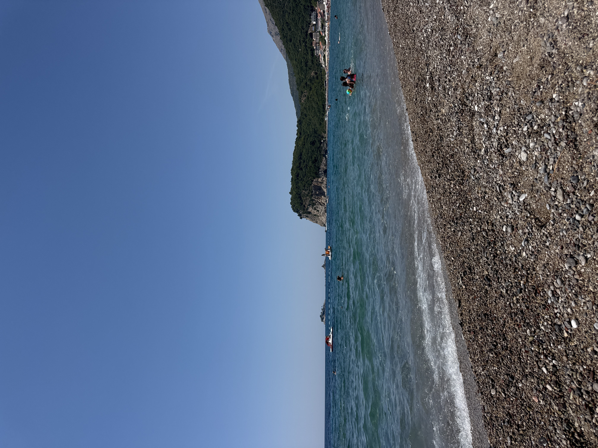 The image depicts a sunny day at a pebble beach in Đurovići, Montenegro. The sky is a clear, bright blue. The foreground is dominated by a dense layer of pebbles, with some small waves gently lapping at the shore. In the mid-ground, the turquoise water is populated by several individuals swimming and enjoying the sea. A red pedal boat with a person on it is visible further out. In the background, a lush green, mountainous coastline rises steeply from the sea. Several buildings are nestled along the base of the mountains, suggesting a settlement. Two small, rocky islands are visible in the distance, with a larger boat or ship anchored between them. The overall scene conveys a serene and idyllic summer beach atmosphere.