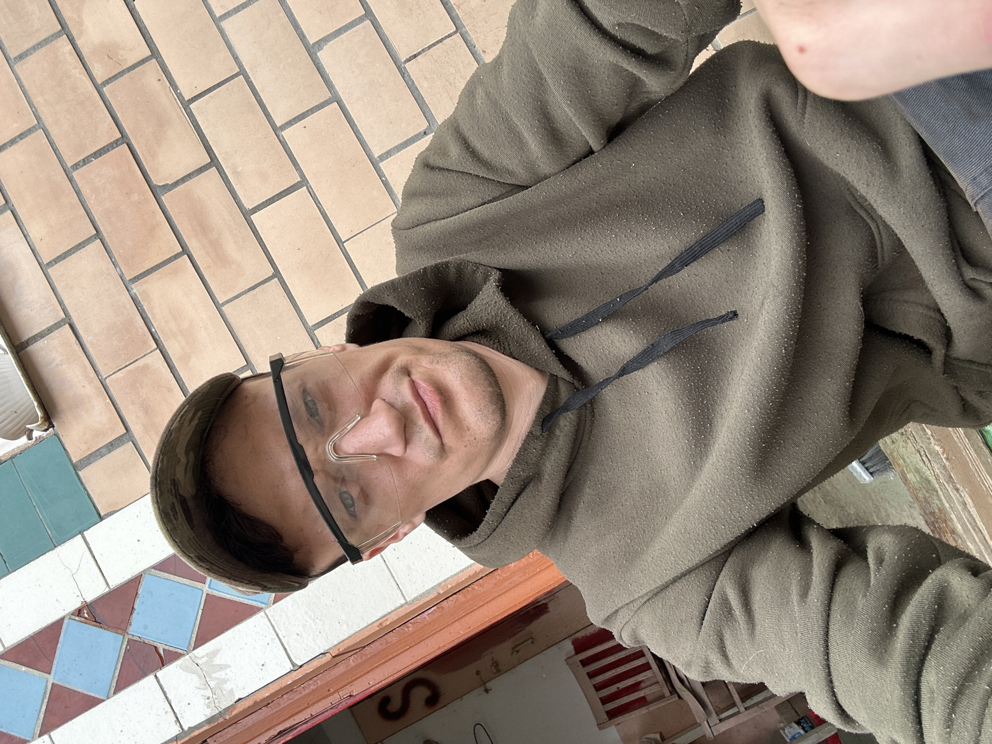In Makiv, Ukraine, a man wearing a camouflage baseball cap and protective goggles is captured in a selfie. He is dressed in a dark green hooded sweatshirt with drawstrings. The background features a brick wall and a decorative tiled surface with diamond patterns in blue, white, and red. An open doorway reveals a glimpse of a chair inside a room. The man is looking directly at the camera with a neutral expression. The overall setting appears to be outdoors, possibly near a building under construction or renovation.