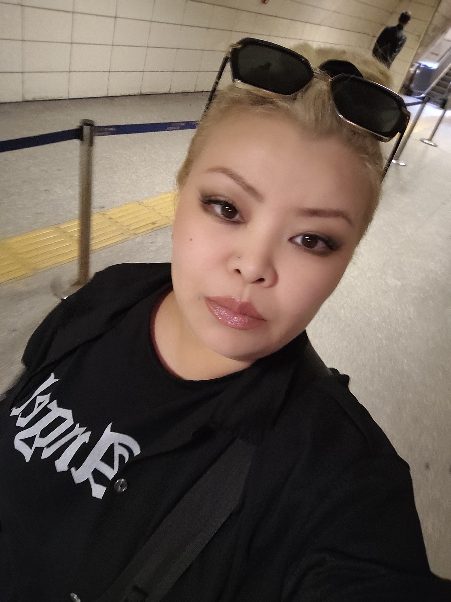 A medium-shot selfie of a woman with light blonde hair and sunglasses resting on her head, taken in an indoor public space, potentially a subway station in Sarıyer, Turkey. The woman is wearing a black t-shirt with white gothic-style lettering and a black jacket. She has a nose piercing. Background includes a yellow tactile paving, a blue railing, white tiled walls, and a blurred figure of a person near an escalator.