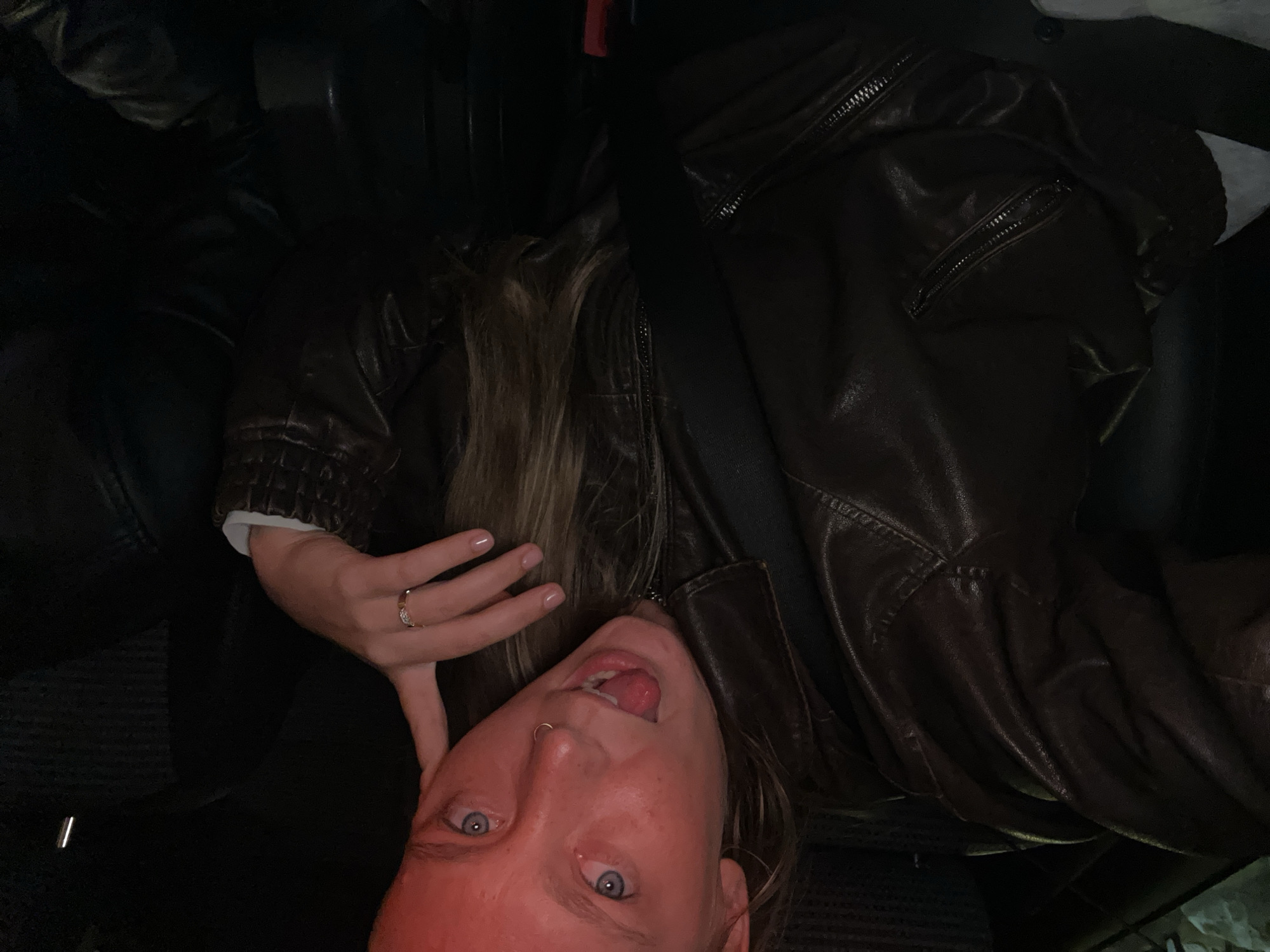 A person with blue eyes and blonde hair is pictured inside a vehicle, likely a car, wearing a dark brown leather jacket and a seatbelt. The individual has a nose piercing and is sticking their tongue out, with their left hand near their head. The interior of the car appears to be dark, with black leather seats. The location in Noisy-le-Grand, France is not discernible from the image.