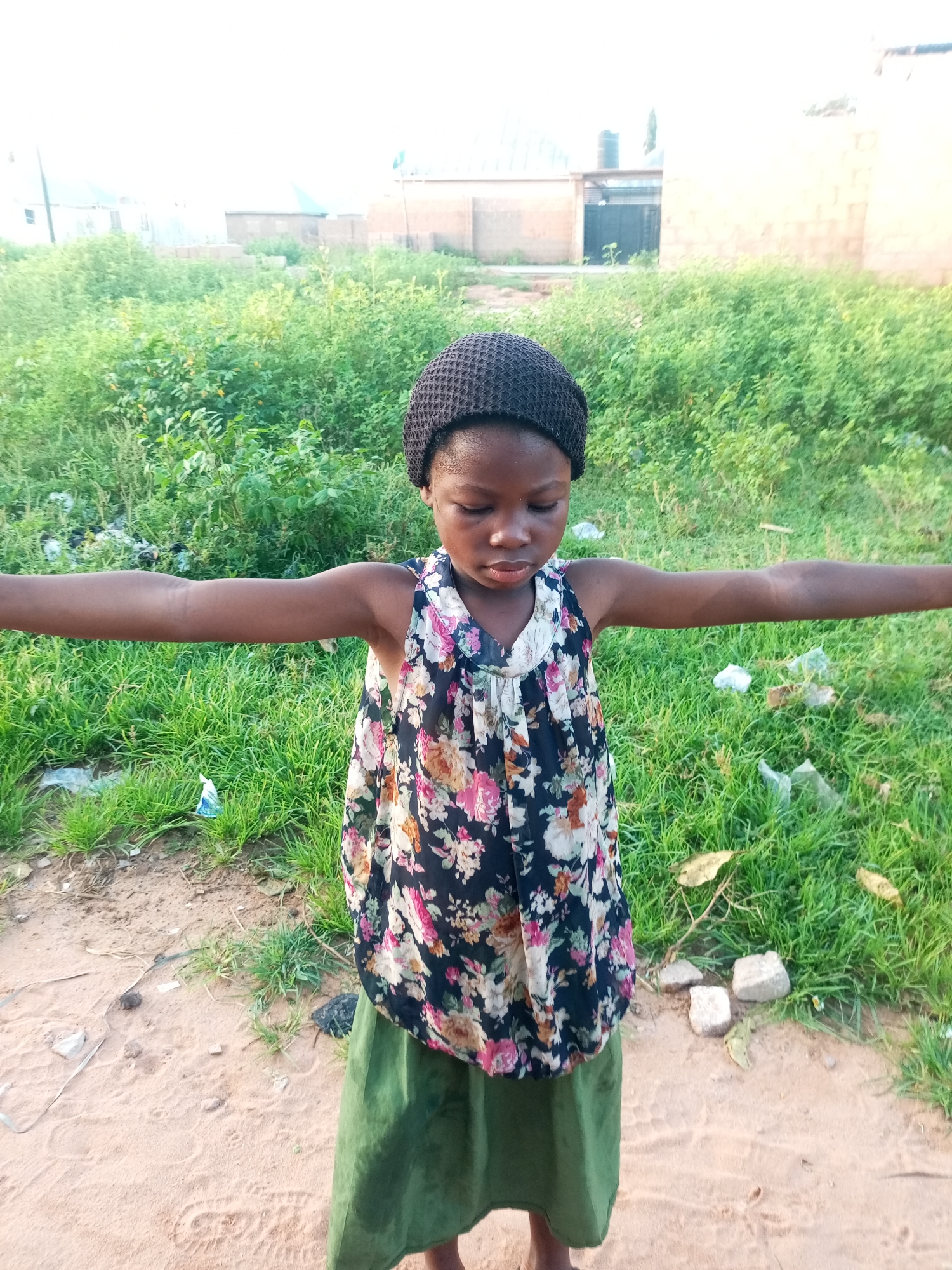 A young Black girl, wearing a dark knitted cap and a floral-patterned dress with a green skirt, stands with her arms outstretched horizontally. She is positioned in the center of the frame, looking downwards with her eyes closed. The background features overgrown green grass, some scattered debris, and distant buildings. The ground in the foreground is sandy with some scattered rocks and leaf litter. The lighting suggests it is daytime, possibly late afternoon.