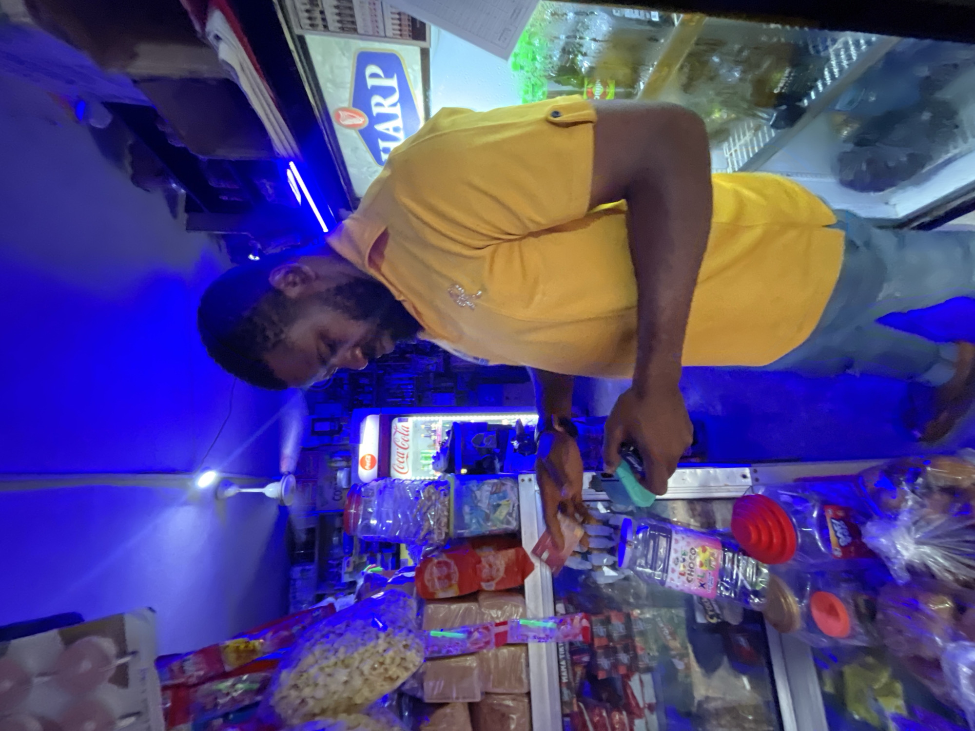 A man in a yellow shirt and jeans is shown at an angle in a retail store in Jos, Nigeria. He is holding money in his left hand. Visible products include popcorn, Coca-Cola, bread, and candy in plastic containers, arranged on shelves. A Sharp sign is visible above a refrigerated display case. The lighting is predominantly blue. A ceiling fan is visible.