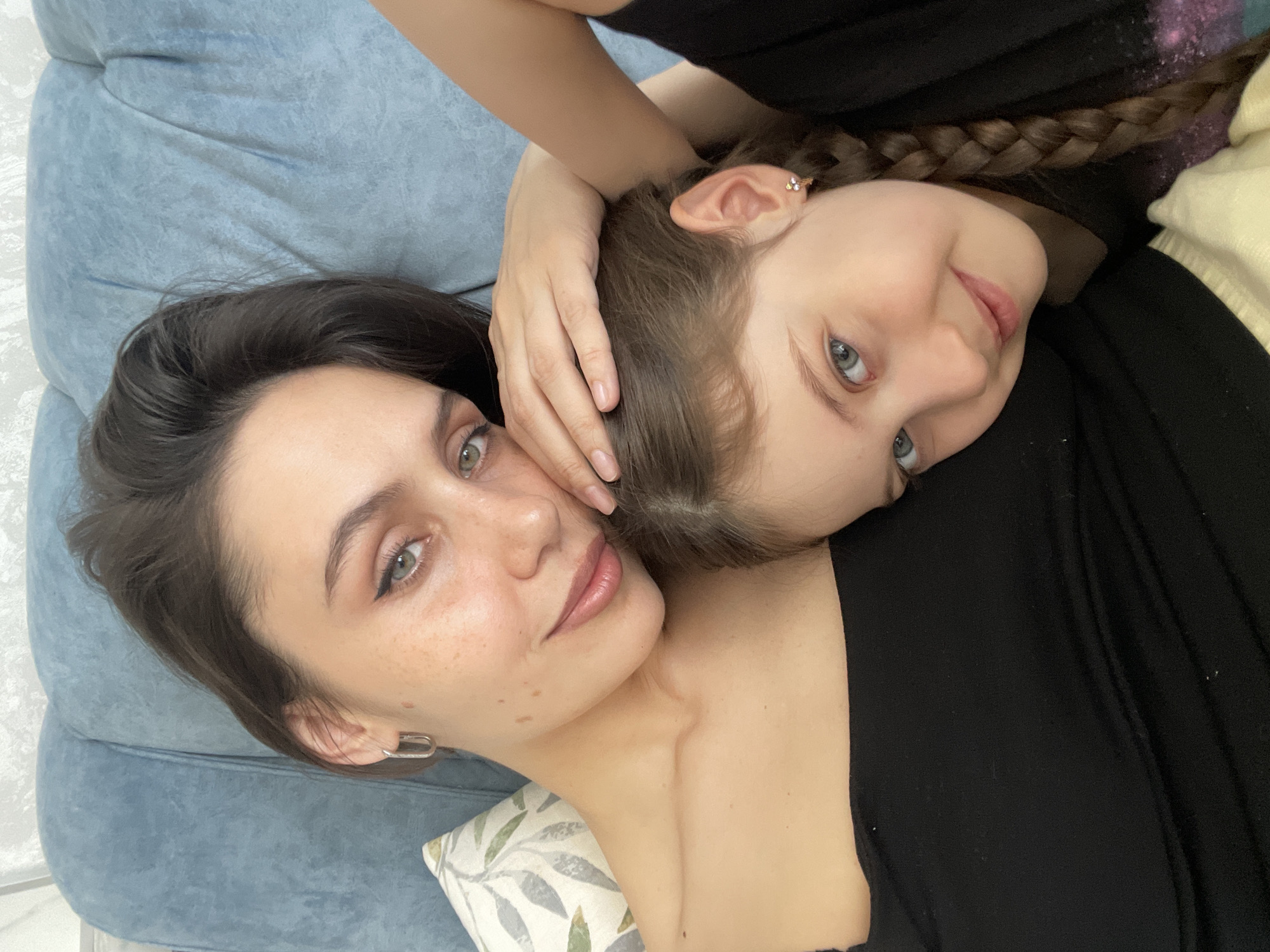 Two women are lying close together. The woman on the left has dark brown hair, light skin with freckles, and wears a silver rectangular earring. The woman on the right has light brown hair braided into a braid and wears a black top. They are lying on a blue patterned surface, possibly a couch. There is a pillow with a leaf pattern visible at the bottom. The location is unknown.
