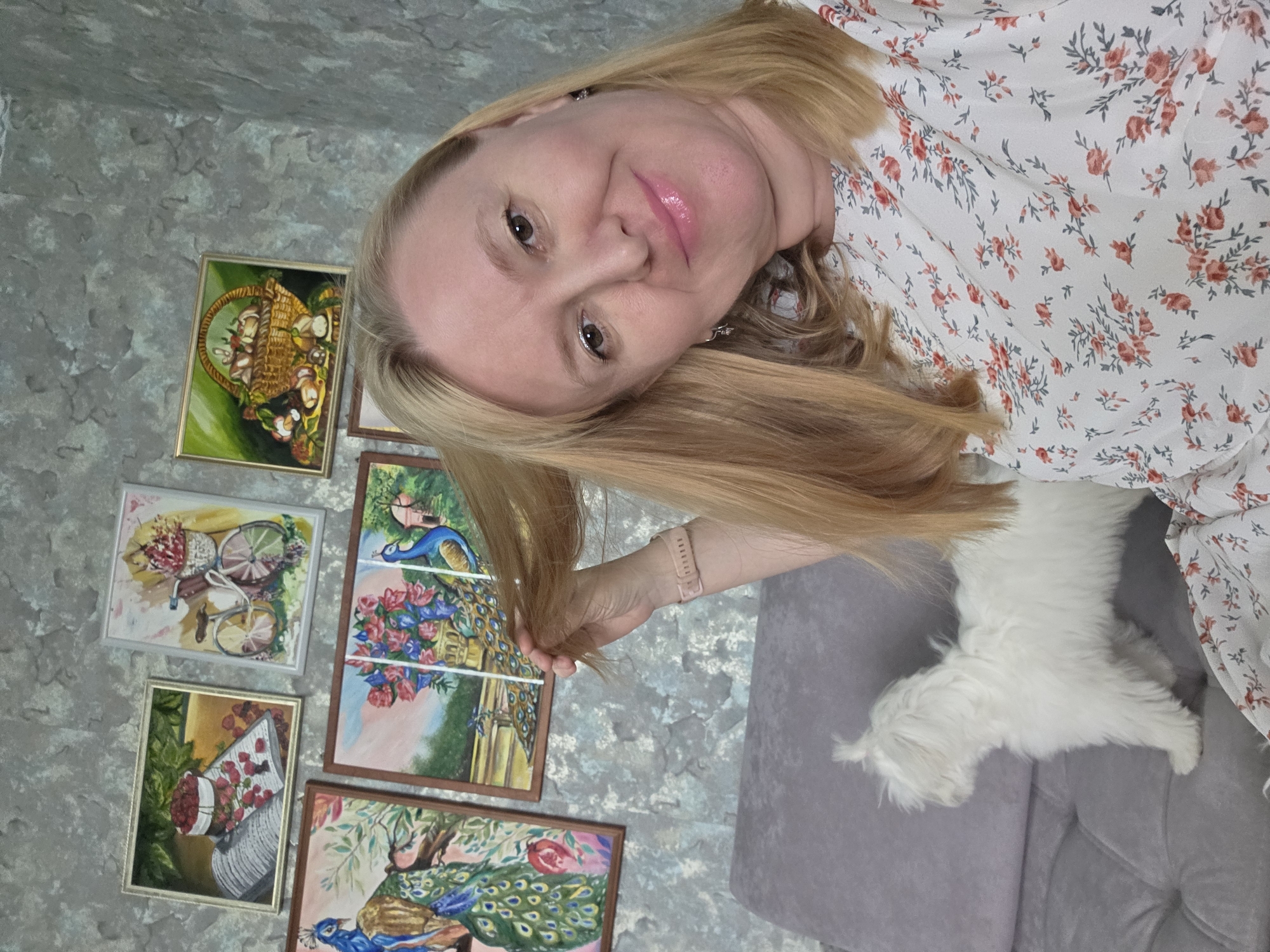 The image depicts a woman with blonde hair and fair skin, wearing a white dress with a floral print. She has a friendly expression and is looking at the camera. To her left, there are several framed paintings on a textured wall. These paintings appear to be of still life and nature scenes, including a basket of mushrooms, a bicycle with flowers, a peacock with a bouquet of flowers, and another peacock with a rose. A small white dog is visible on a grey sofa to the woman's right, also looking towards the camera. The overall scene suggests a home environment in Novocheboksarsk, Russia. The lighting appears to be indoor, and there are no specific indicators of time of day or weather. The woman's pose and smile suggest a casual and happy moment, possibly a selfie.