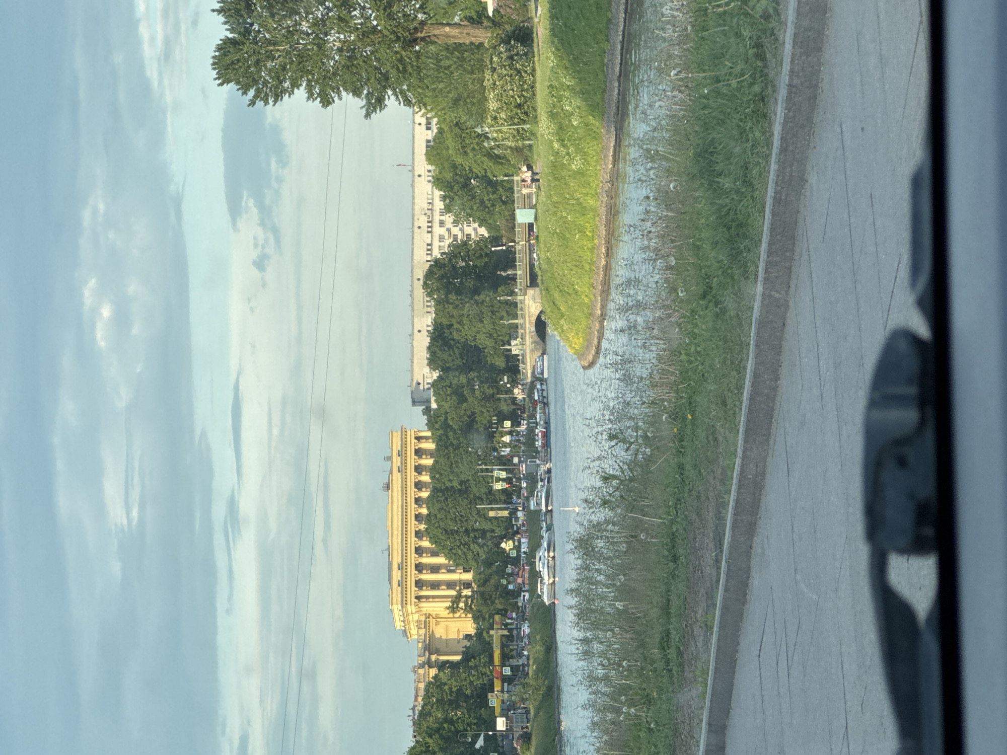 The image captures a vibrant urban scene in Saint Petersburg, Russia, under a bright, cloudy daytime sky. The perspective suggests it was taken from a vehicle, with a dark, blurred element partially visible on the extreme right foreground.
A calm body of water, likely a river or canal, stretches horizontally across the middle ground. On the far side of the water, a bustling city street is visible, flanked by a row of buildings and lush green trees. A prominent, light-colored building with intricate classical architecture, featuring multiple windows and decorative elements, stands out among the structures. Further along, other multi-story buildings contribute to the urban skyline. The street is busy with numerous cars, indicating active traffic; some vehicles appear to be stopped. Several small, square road signs are visible along the street, but their text is too distant to read.
On the near side of the water, a grassy bank slopes up to a paved walkway or path, bordered by a concrete curb. Thin power lines or cables are faintly visible against the sky, especially in the upper right portion of the image. A few indistinct figures of people can be seen on the far side, near the buildings or by the water's edge, adding to the sense of everyday city life. The overall impression is one of a typical, active day in an architecturally rich European city.