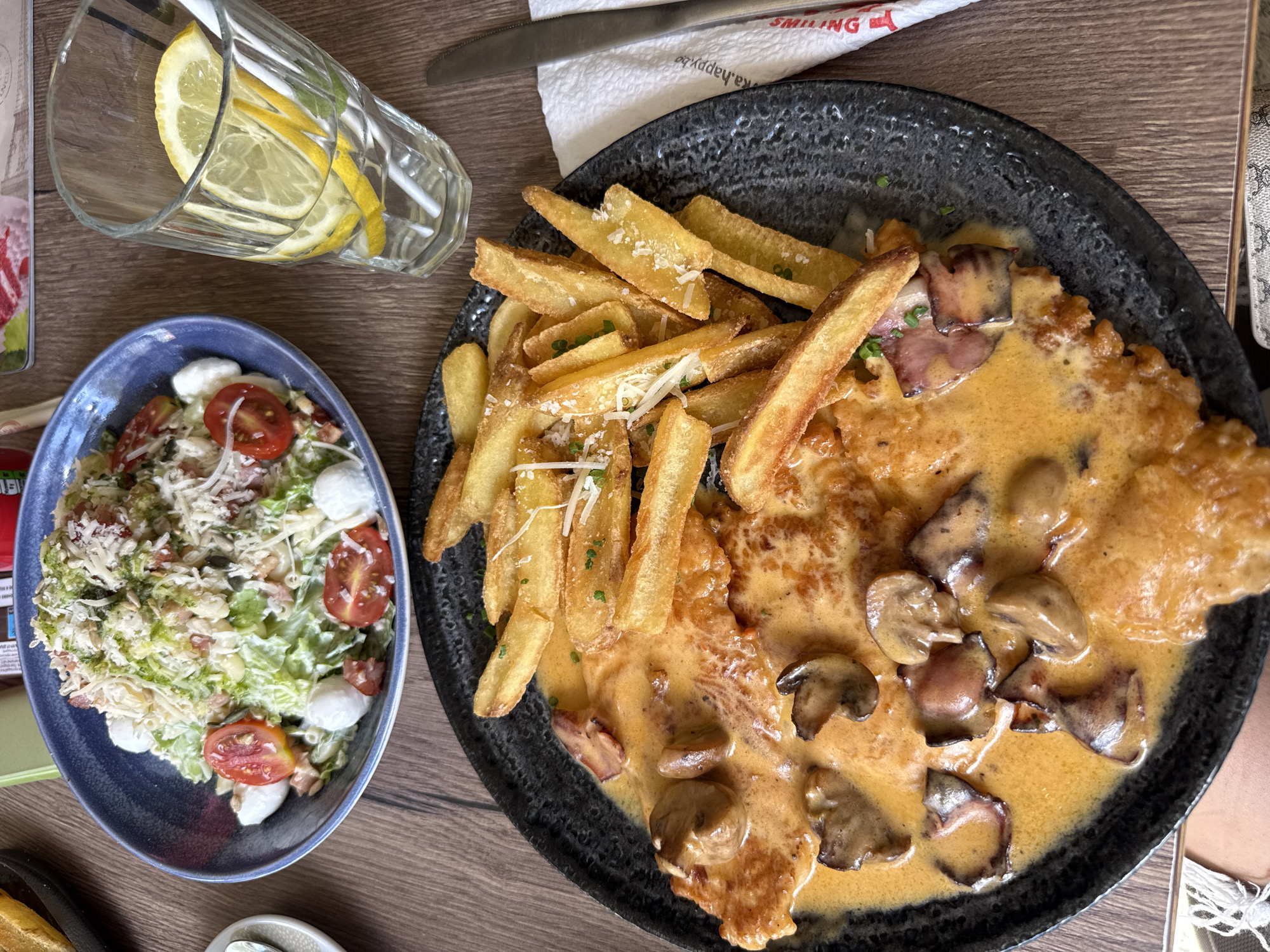 The image shows a table at a restaurant, likely in Plovdiv, Bulgaria, given the context clues like European cuisine and potential location of the restaurant. A large, dark, textured plate dominates the right side of the image. It contains a schnitzel covered in a creamy, mushroom sauce, accompanied by a serving of fries sprinkled with herbs and grated cheese. To the left is a blue bowl containing a salad with lettuce, tomatoes, mozzarella balls, and grated cheese. A glass of water with lemon slices stands upright near the salad. In the background, a knife rests on a white napkin, and sections of table decorations are partially visible. The table has a wooden surface.