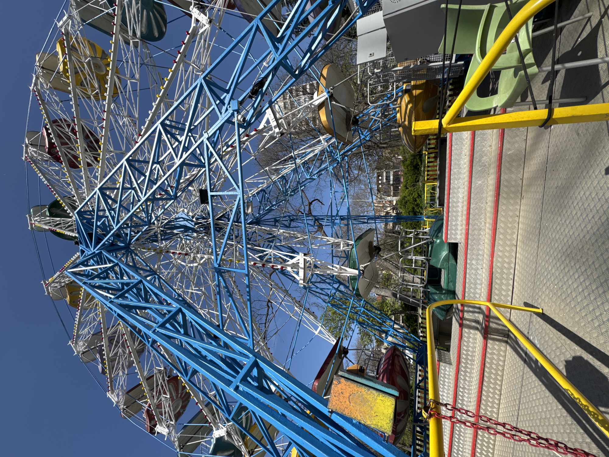 The image displays a vibrant Ferris wheel located in an amusement park or fairground in Varna, Bulgaria, captured on a clear, sunny day. The dominant structure of the Ferris wheel features an intricate lattice of blue and white metal girders and spokes. Attached to the wheel are numerous passenger gondolas or cabins, painted in bright primary colors such as yellow, red, and green. Small lights are embedded along the framework of the wheel, indicating its operation during evening hours.
In the foreground, a durable metal platform with a diamond-plate surface serves as the boarding area for the ride. Red stripes are painted onto the platform, likely for guidance, and sturdy yellow metal handrails are installed for safety. A red metal chain is stretched across a section of the handrail, suggesting a temporary closure or restricted access point. A few light green plastic chairs are also visible near the platform, possibly for a waiting area.
The background shows a brilliant blue sky, reinforcing the clear weather. Beyond the mechanical structures, there are trees, some still bare and others just beginning to sprout green leaves, along with glimpses of light-colored buildings in the distance. The absence of people gives the scene a quiet, almost deserted atmosphere, possibly indicating that the photo was taken before opening hours or during an off-peak period. No visible text is present in the image.