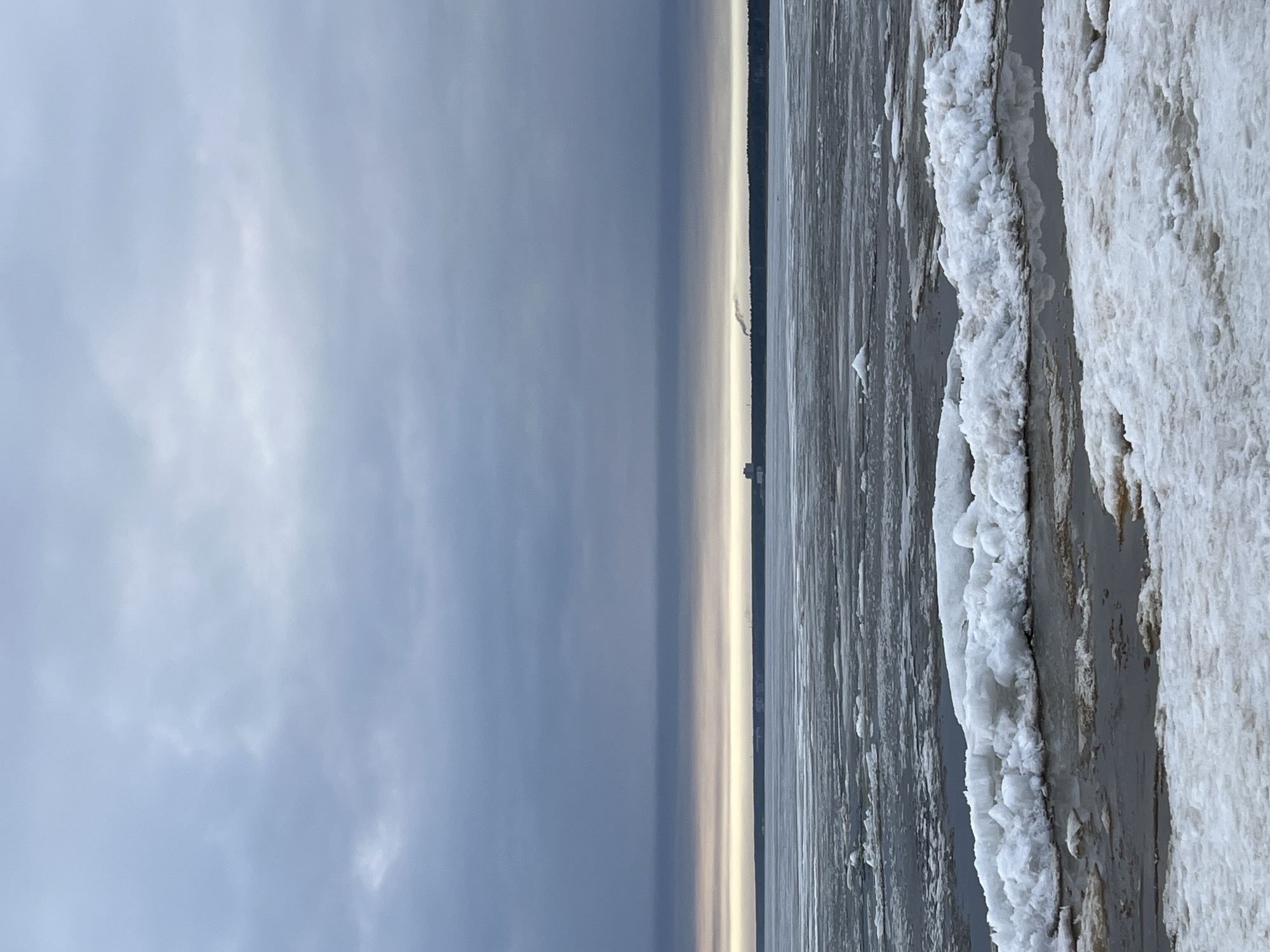 The image shows a winter scene in Sestroretsk, Russia. The sky is overcast with grey clouds. To the right, the shoreline is covered in ice and snow, with textured formations suggesting layers of accumulation. The water is partially frozen, displaying a mix of open water and ice patches. In the distance, a faint outline of a structure is visible on the horizon.