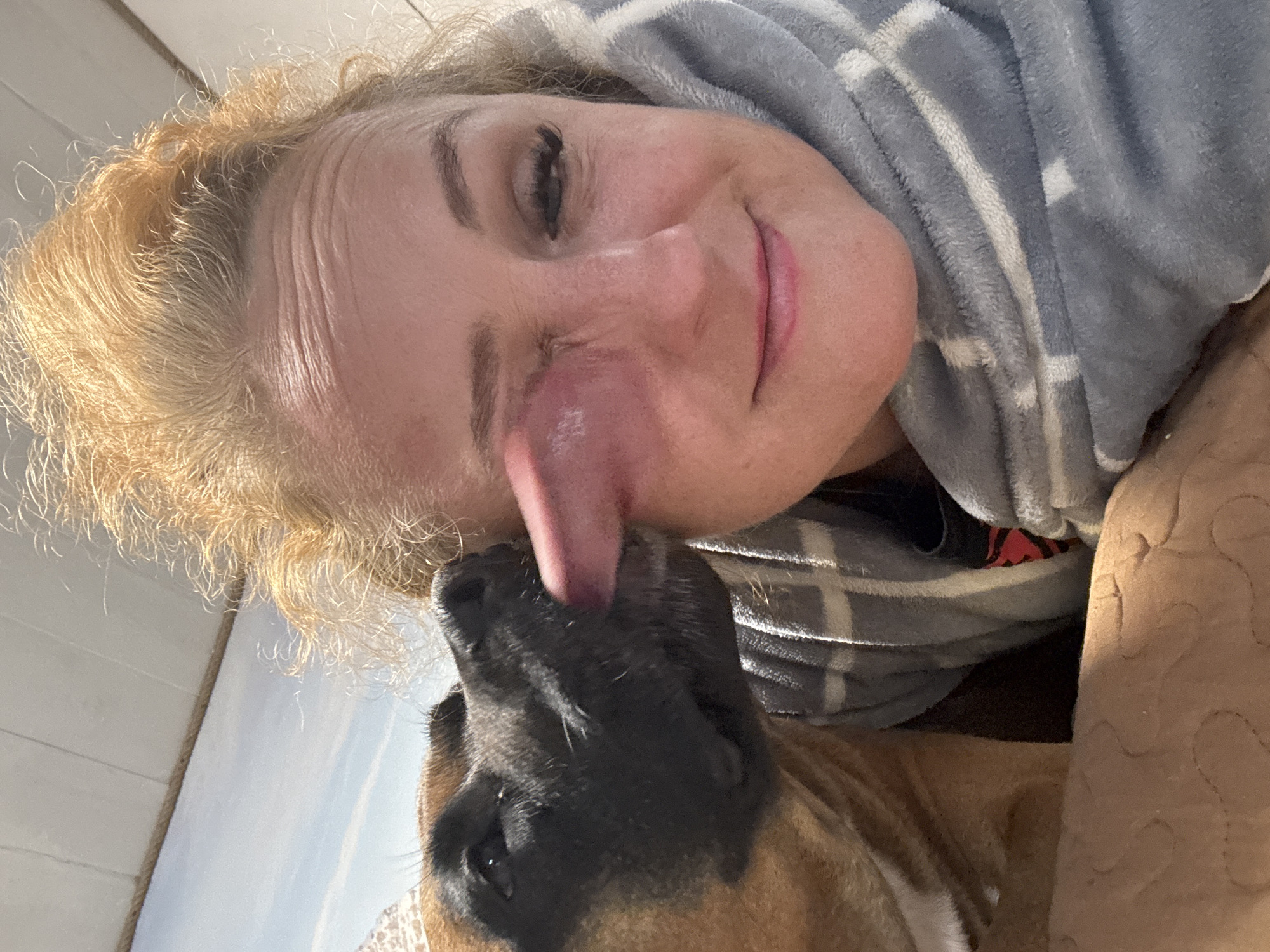 An indoor, eye-level shot captures a close, affectionate interaction between a woman and a dog. The image is oriented with the woman's head and the dog's head appearing to be on their sides, suggesting they are lying down.
The woman is visible from her upper chest to the top of her head. She has fair skin, light blonde or light brown curly hair, and is wearing what appears to be a grey and white striped fuzzy blanket or robe draped over her upper body. Her right eye is visible and closed, with dark eyelashes. She has a gentle smile on her lips, indicating happiness and contentment.
A dog, with a dark muzzle and brown fur, is positioned close to the woman's face. The dog's pink tongue is extended and actively licking the woman's cheek and nose area, which appears slightly reddened from the contact. The dog's eye is partially visible, looking upwards towards the woman.
The background includes a light-colored wall or paneling and a light brown, textured surface, possibly another blanket or piece of furniture. The lighting is bright and natural-looking, suggesting it is daytime. The overall mood of the image is warm and loving, centered around the bond between the woman and her pet. No text is visible in the image.