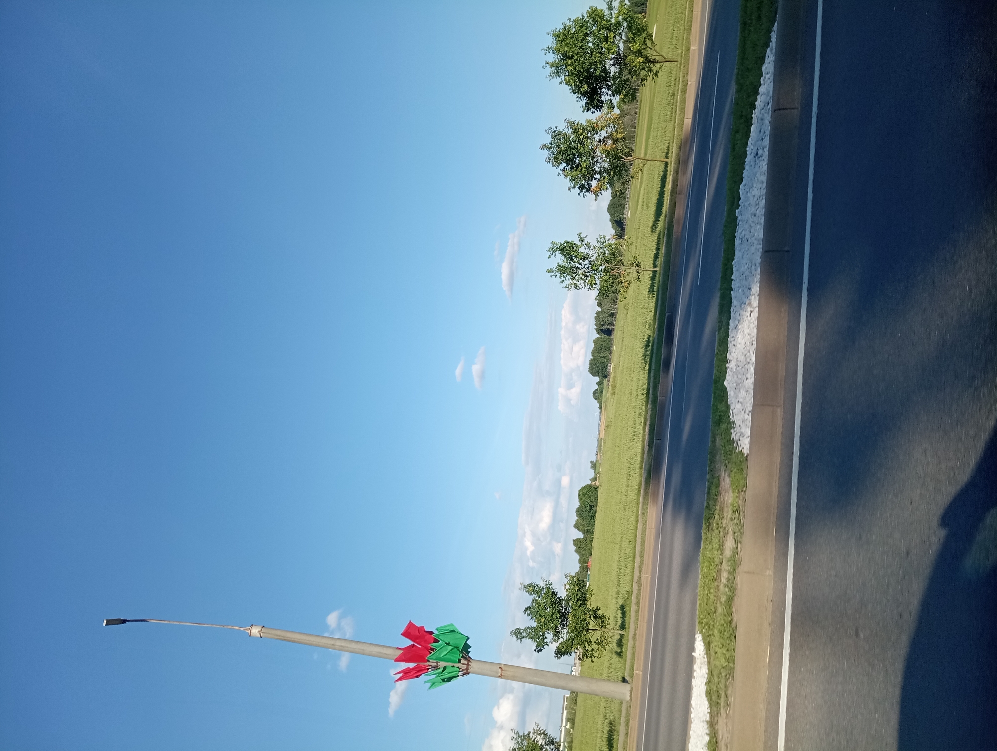 An outdoor, daytime, low-angle, wide shot captures a scene on the outskirts of Буйніцкі сельскі Савет, Belarus. The sky is a clear, bright blue with a few scattered white clouds. In the foreground, a dark asphalt road runs horizontally across the bottom of the frame, with a white stripe marking the lane. Adjacent to the road is a curb and a strip of grass with white gravel. Beyond the grass, a green field stretches into the distance, dotted with several young trees and a line of mature trees on the horizon. A light pole stands prominently on the left side of the image, its metallic surface weathered. Attached to the pole, about a third of the way up, is a cluster of red and green flags arranged in a circular pattern. The flags appear to be made of fabric and are gently waving. A small tree is positioned in front of the light pole, its leaves a vibrant green. The overall impression is a rural or semi-rural landscape under a clear sky.