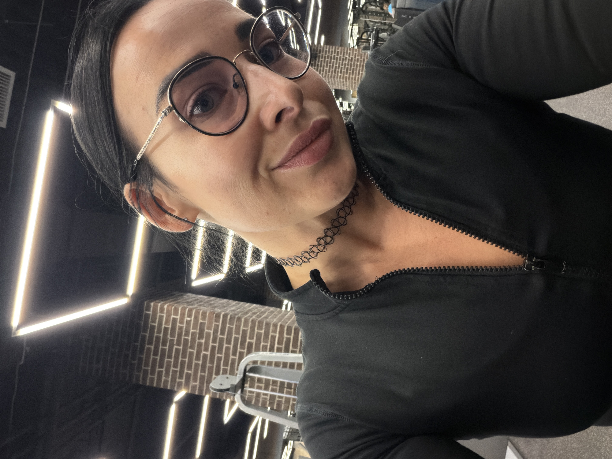 A selfie captures a woman from the chest up, positioned at an angle, in what appears to be a gym or fitness studio in Saint Petersburg, Russia. She has dark hair, styled neatly, and wears round, framed glasses. Her lips are slightly parted, revealing a neutral to gentle expression. Around her neck is a black, intricate choker necklace. She is dressed in a black top with a zipper that is partially unzipped.
The background features dark walls illuminated by bright, linear LED lights arranged in geometric patterns. A section of the wall behind her is adorned with light brown brickwork. The lighting suggests an indoor setting, possibly during the evening or in a windowless space. No specific text is visible in the image.