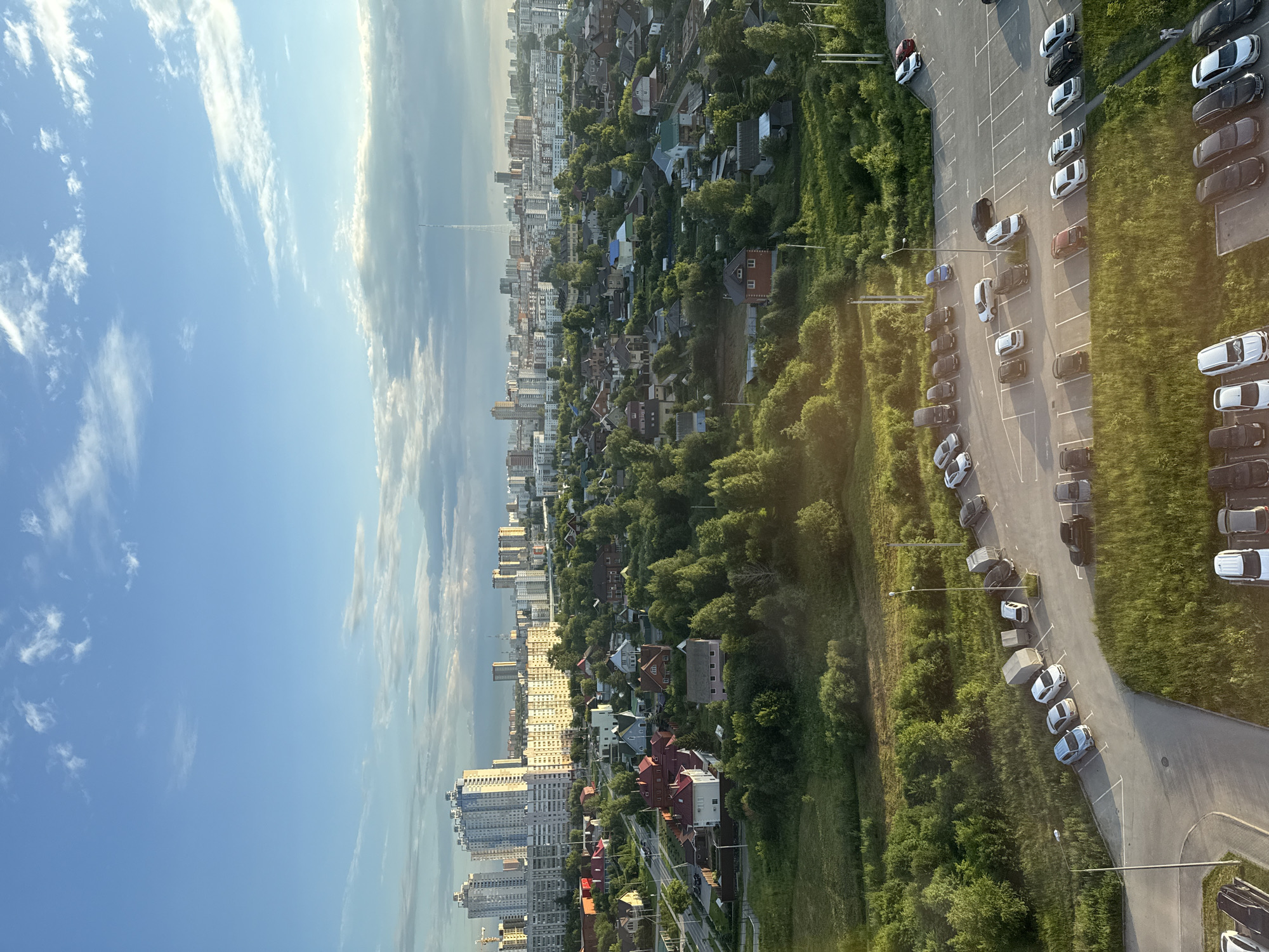 This is an aerial, landscape-oriented photograph taken from a high vantage point, likely a building or drone, overlooking a residential and urban fringe area. The image is vertically oriented, with the sky occupying the left half and the land on the right.
The scene depicts a sprawling cityscape transitioning into a densely wooded area dotted with houses and then giving way to a parking lot filled with cars. In the distance, tall, modern apartment buildings form a prominent skyline. Closer to the foreground, there is a neighborhood of smaller houses with red and white roofs, nestled amongst lush green trees. A paved road winds through this area, with cars parked along its sides and in designated parking spots. The parking lot on the far right is filled with numerous vehicles of various colors and models.
The time of day appears to be late afternoon or early evening, indicated by the warm, golden sunlight casting long shadows and highlighting the green foliage. The sky is a clear blue with scattered white, wispy clouds, suggesting fair weather.
While no people are clearly discernible, the presence of numerous parked cars and residential buildings indicates human activity and habitation. The overall mood is peaceful and serene, capturing a typical suburban or semi-urban environment bathed in the soft light of the setting sun. There is no visible text in the image. The location context provided is Perm, Russia.