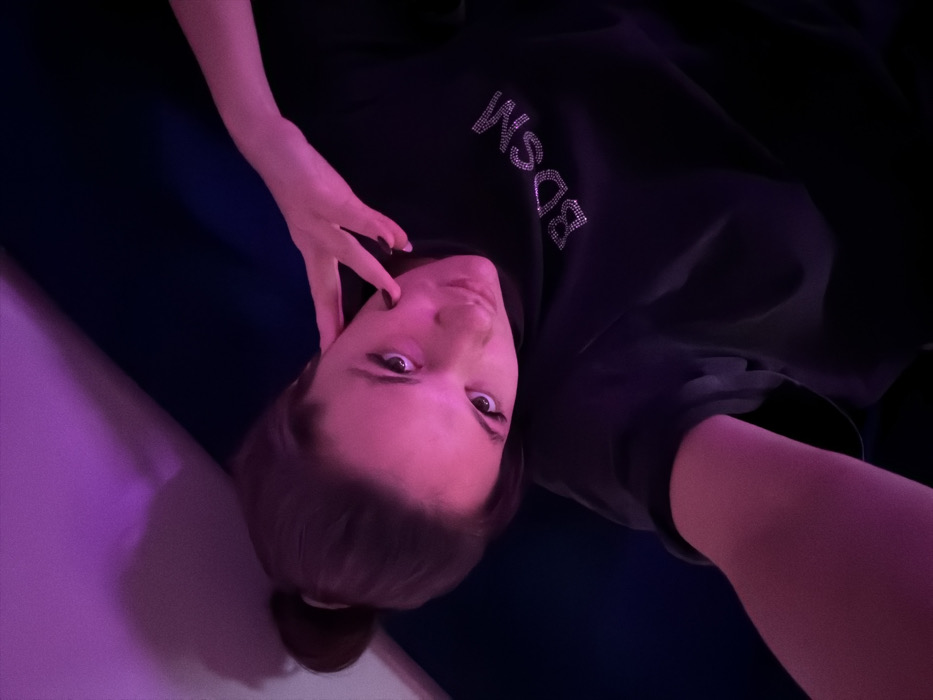 A female individual is depicted indoors, captured in a selfie at an elevated angle. Her dark hair is styled in a bun, and she wears a dark-toned long-sleeved shirt featuring 'BDSM' spelled out in rhinestone-like embellishments on the chest. Her left hand, with dark-painted nails, rests with fingers against her left cheek and neck, while her right arm extends forward, presumably holding the camera. She gazes directly into the lens. The background shows a dark blue surface, likely furniture, and a lighter wall segment, all illuminated by strong purple-pink ambient lighting. The indoor setting lacks any visual cues to determine its specific location within Belgrade, Serbia.