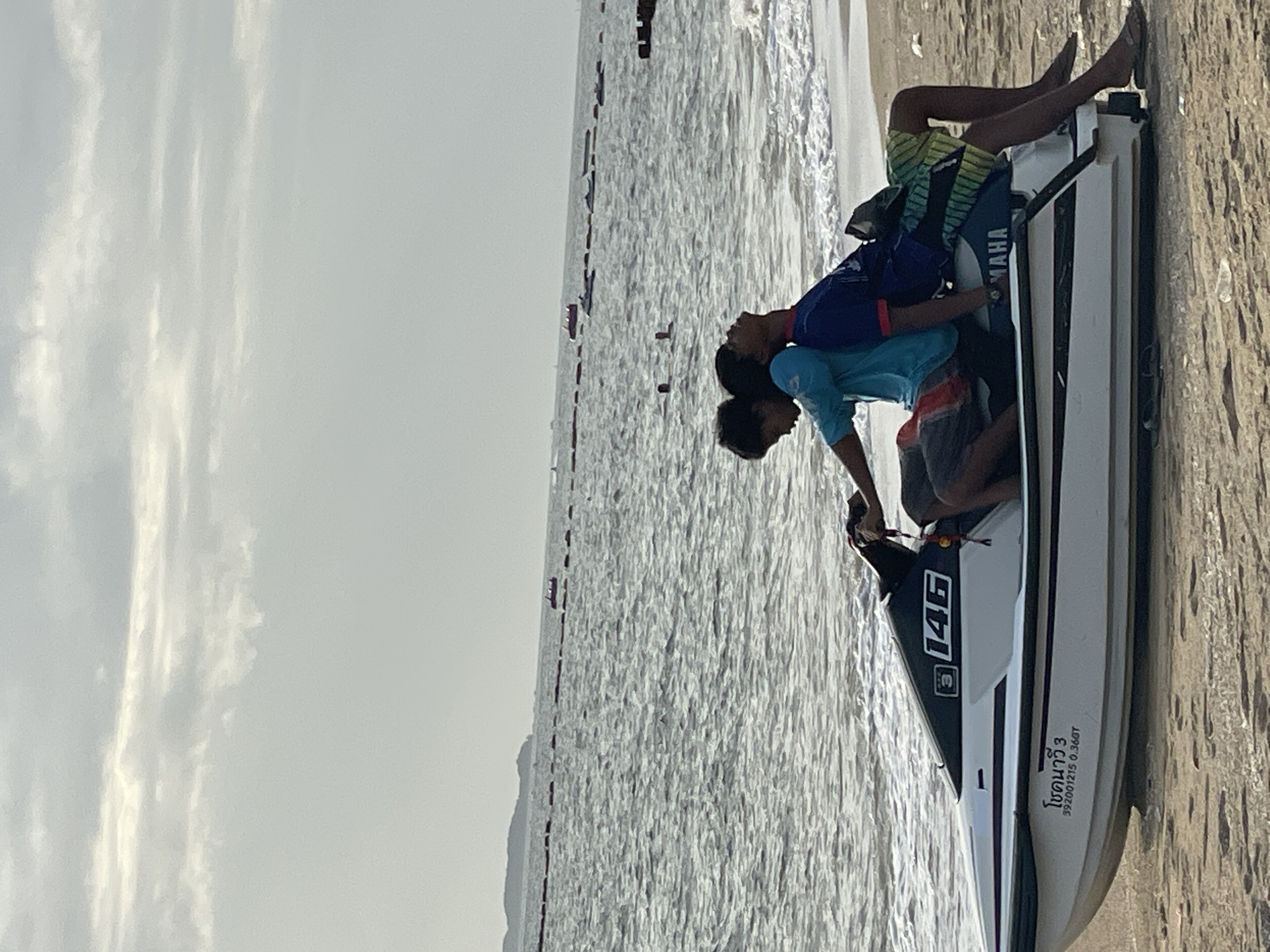 Two young men are sitting on a white and black Yamaha jet ski on the sandy beach of Pattaya City, Thailand. The jet ski is positioned on the shore with the ocean in the background. The man in the front is wearing a blue t-shirt and patterned shorts, and the man behind him is wearing a dark blue t-shirt with red accents and green and black patterned shorts. Both are looking towards the water. The sky is overcast with clouds. In the distance, the ocean is visible with small waves lapping the shore. There are several other jet skis and a small boat further out in the water. A line of buoys stretches across the water in the mid-ground. The beach is sandy with some scattered debris.