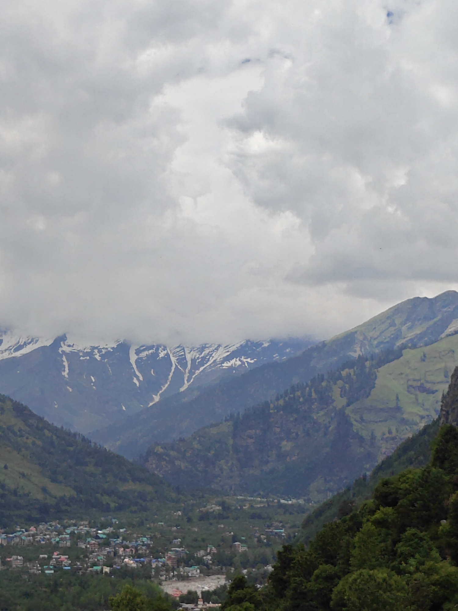 The image depicts a panoramic view of the town of Vashisht, India, nestled within a valley in the Himalayan mountains. The sky is overcast with dense gray clouds. In the foreground, lush green trees frame the lower right portion of the scene. Beyond the trees, a town with buildings of varying colors and sizes is spread across the valley floor. A river or body of water is visible running through the town. The middle ground is characterized by rolling green hills dotted with trees. In the background, imposing mountains rise, their peaks partially covered with snow and shrouded in mist or low-lying clouds. The overall impression is one of a scenic, mountainous landscape with a densely populated town in the valley.