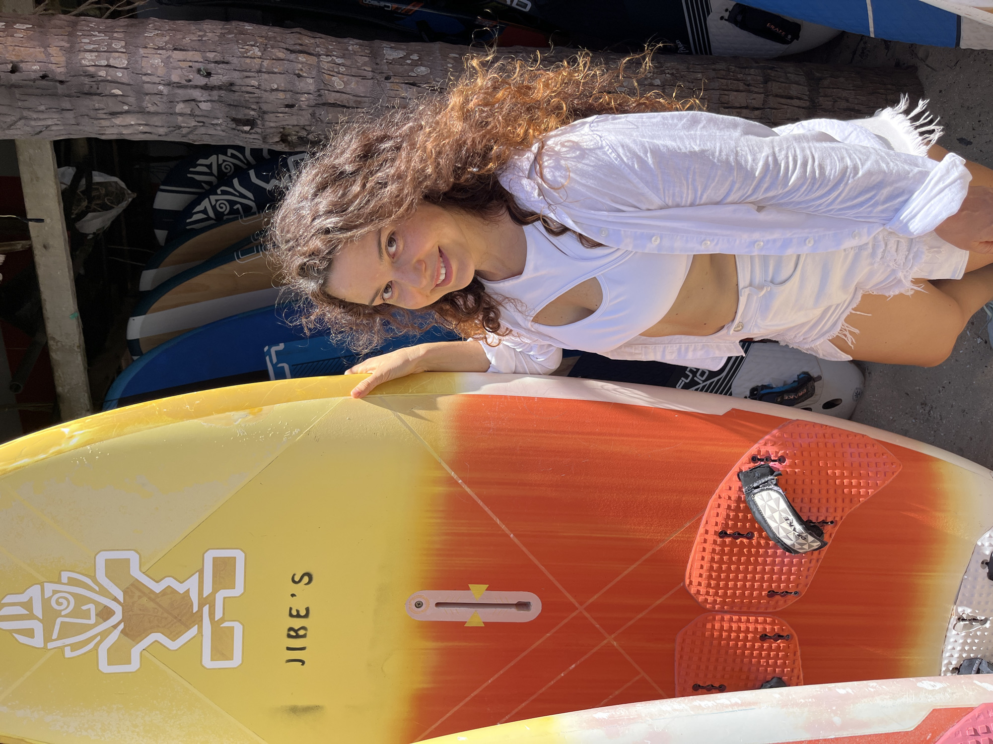 A woman with long, curly brown hair and fair skin stands next to a yellow and orange surfboard. She is wearing a white crop top with a cutout at the chest, a white open-buttoned shirt, and distressed white denim shorts. The surfboard has the text "JIBE'S" printed on it in black, and a graphic of a stylized face at the top. Behind the woman, several other surfboards are leaning against a wooden structure, and a large palm tree trunk is visible to her right. The scene appears to be outdoors, likely near a beach or a surf shop, given the presence of the surfboards and the palm tree. The ground is covered with sand or gravel. The overall lighting suggests a sunny day. The location is Phan Thiết, Vietnam.