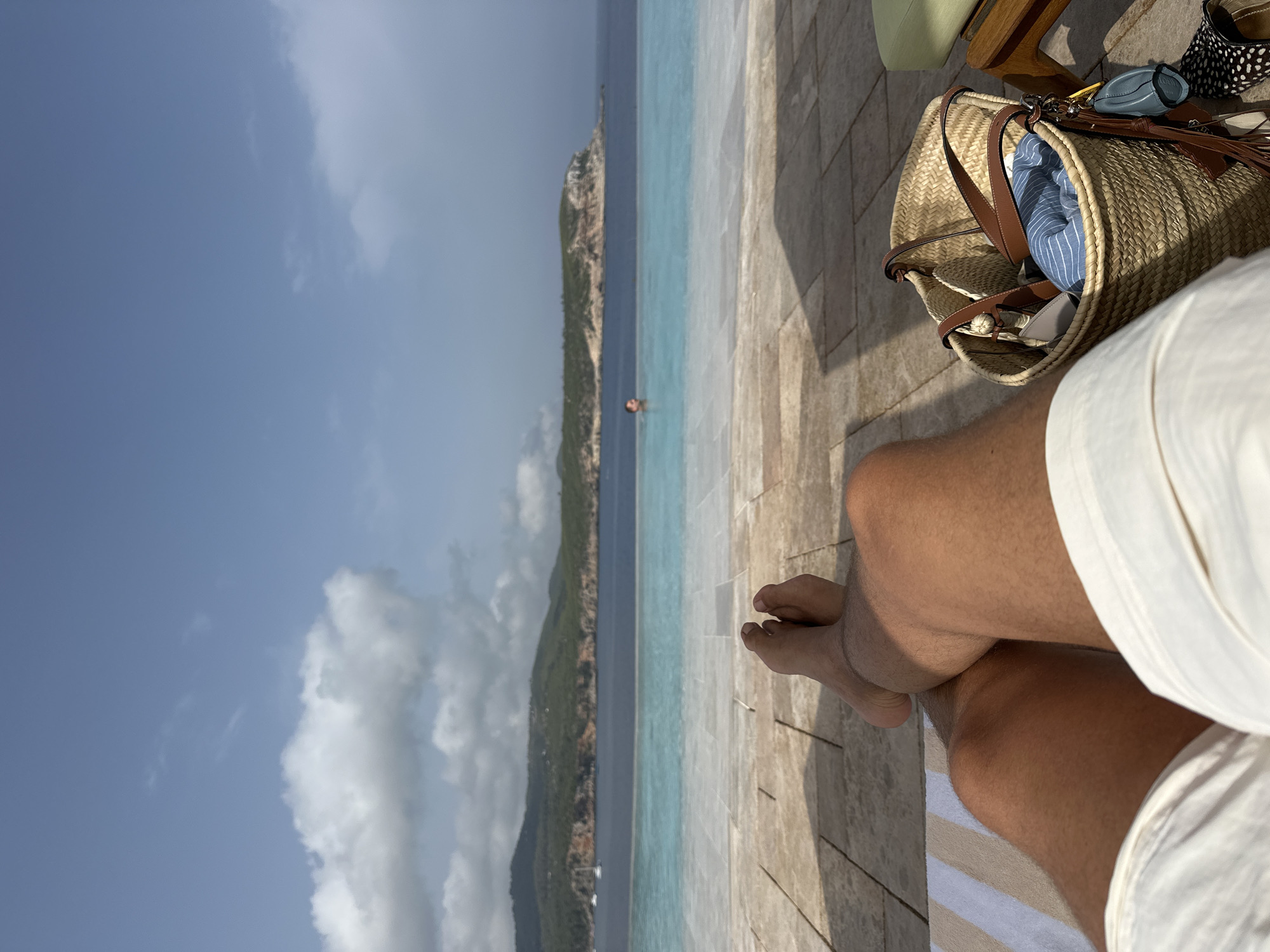 In a first-person perspective, the image shows a person relaxing on a lounge chair near an infinity pool overlooking the sea. The person's legs and feet are visible in the foreground, with white shorts and tanned skin. A straw beach bag with a blue and white striped towel, a small gray pouch, and a blue keychain is placed to the right of the person. In the middle ground, the infinity pool stretches out, meeting the calm blue sea. In the background, a lush green island with a rocky coastline is visible, with a few sailboats anchored in the water. The sky is partly cloudy, with a mix of blue sky and white cumulus clouds. The scene suggests a tranquil vacation setting.