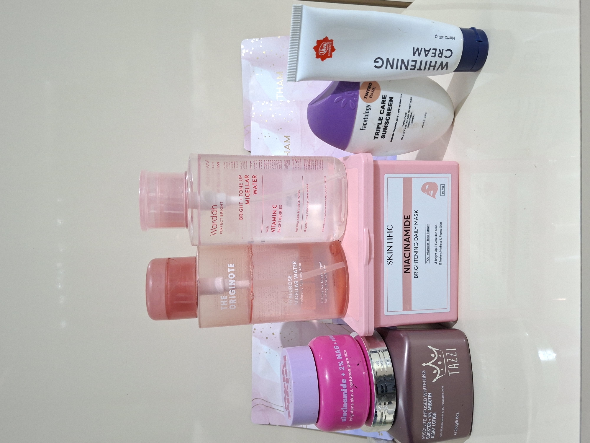 The image presents a collection of skincare products arranged on a flat, reflective surface, likely a table or countertop.
The items include:
* A tube of Viva Cosmetics Whitening Cream (Netto 40g).
* Facetology Triple Care Sunscreen (Tinted Bare, 40ml/1.3oz).
* Two bottles of micellar water: one Wardah Perfect Bright Bright + Tone Up Micellar Water and one The Originote Hyalurose Micellar Water.
* A pink box of Skintific Niacinamide Brightening Daily Mask (30 pcs).
* A pink jar of a skincare product labeled "niacinamide + 2% NAG".
* A brown jar of Tazzi Absolute Infused Whitening Booster + 3% Arbutin Night Lotion (e250g/8.8oz).
* A sheet mask.
The products are primarily white, pink, and brown. The setting is plain and there is no information regarding the location in Kutaglumpang, Indonesia.