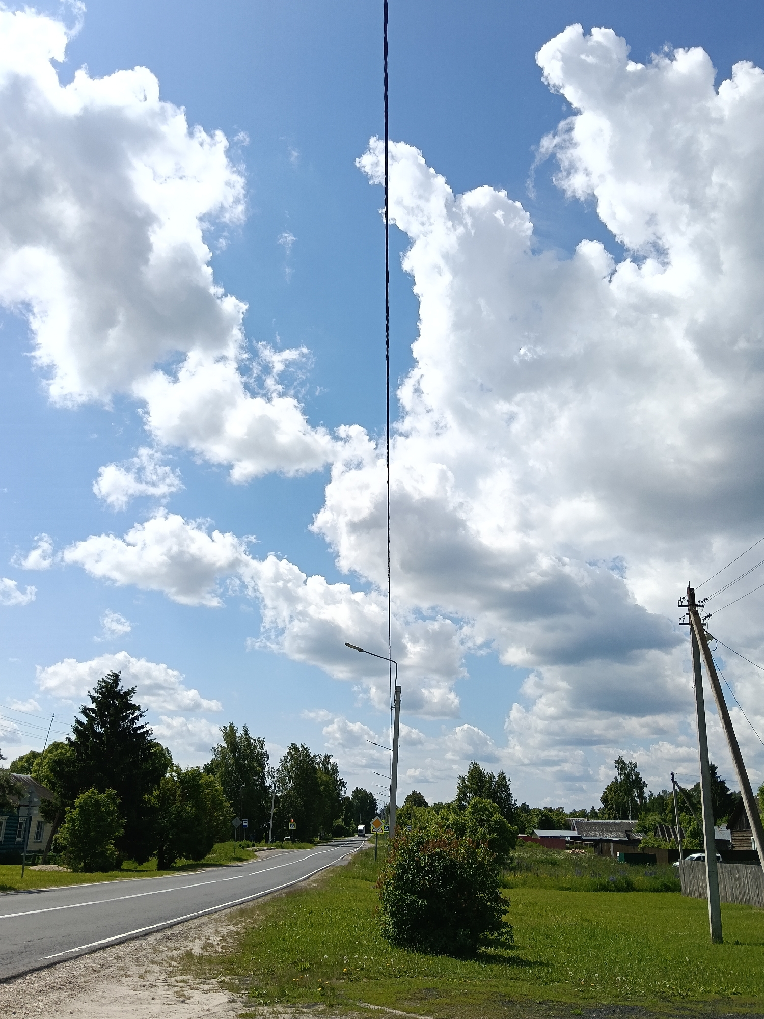 This image depicts a sunny daytime scene in Красный Маяк, Russia, characterized by a rural or semi-urban road winding through a landscape of green vegetation and scattered buildings. The sky is a prominent feature, filled with large, fluffy white cumulus clouds against a clear blue background, indicating fair weather. A tall, dark vertical line, likely a utility wire or cable, stretches from the top to the middle of the frame.
A paved road with white line markings curves gently to the right, disappearing into the distance. Lush green grass lines the roadside, dotted with smaller bushes and trees. On the left side of the road, a dense, dark green evergreen tree stands out, partially obscuring a light blue wooden house. Further down the road on the left, more deciduous trees and utility poles are visible.
To the right of the road, a well-formed bush with reddish-green leaves occupies the grassy verge. A sturdy wooden power pole with several crossarms and wires stands prominently on the right, extending upwards towards the sky. A metal street light pole is also visible further down the road on the right. In the background, beyond the trees, several houses with dark roofs and wooden fences are visible, suggesting a residential area. Road signs are present, including one depicting a pedestrian crossing symbol and another possibly indicating a bus stop. No people or specific activities are visible, conveying a calm and serene atmosphere.