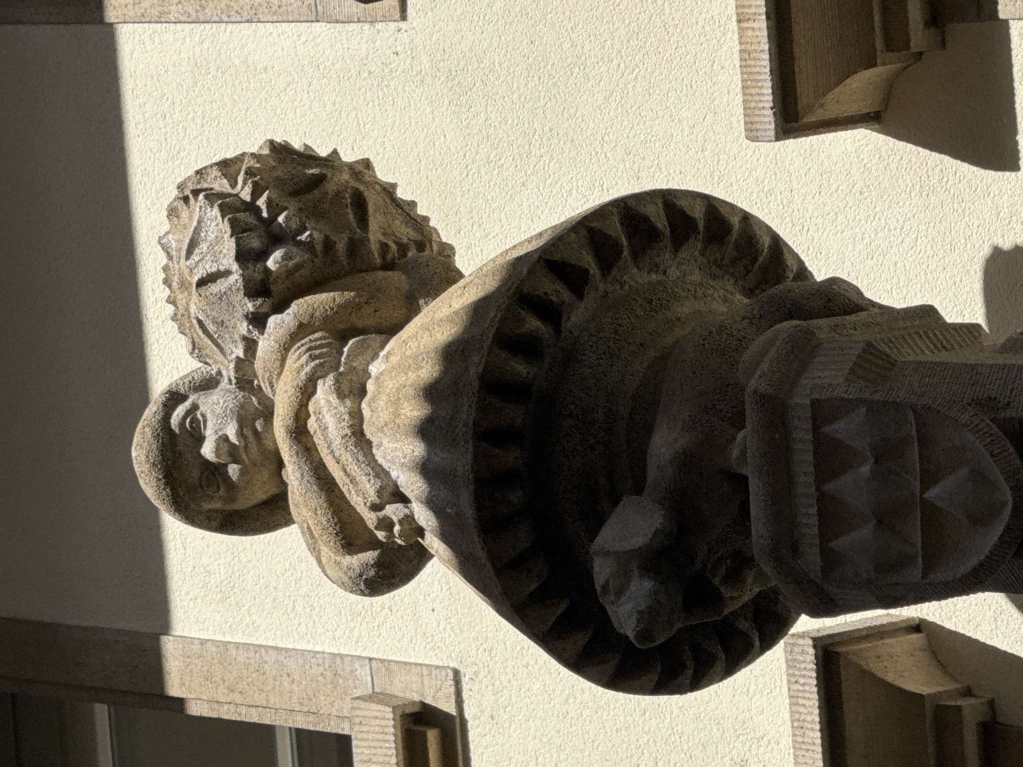 A stone sculpture of a figure wearing a hat and holding a large, spiky object is mounted on a building in Lucerne, Switzerland. The sculpture is located on the facade of a building. The figure is depicted from the chest up, with its arms wrapped around the spiky object. The object appears to be a cluster of sharp, angular protrusions. The figure's face is visible, with a neutral expression. Below the figure is a round, decorative element with triangular protrusions. Further down, a shield-like shape is visible, adorned with a geometric pattern of triangles. The building's facade is a textured yellow wall, with shadows cast by sunlight. Architectural elements, including window frames and ledges, are visible to the left and right of the sculpture. The overall impression is of an ornate architectural detail integrated into the building's exterior.