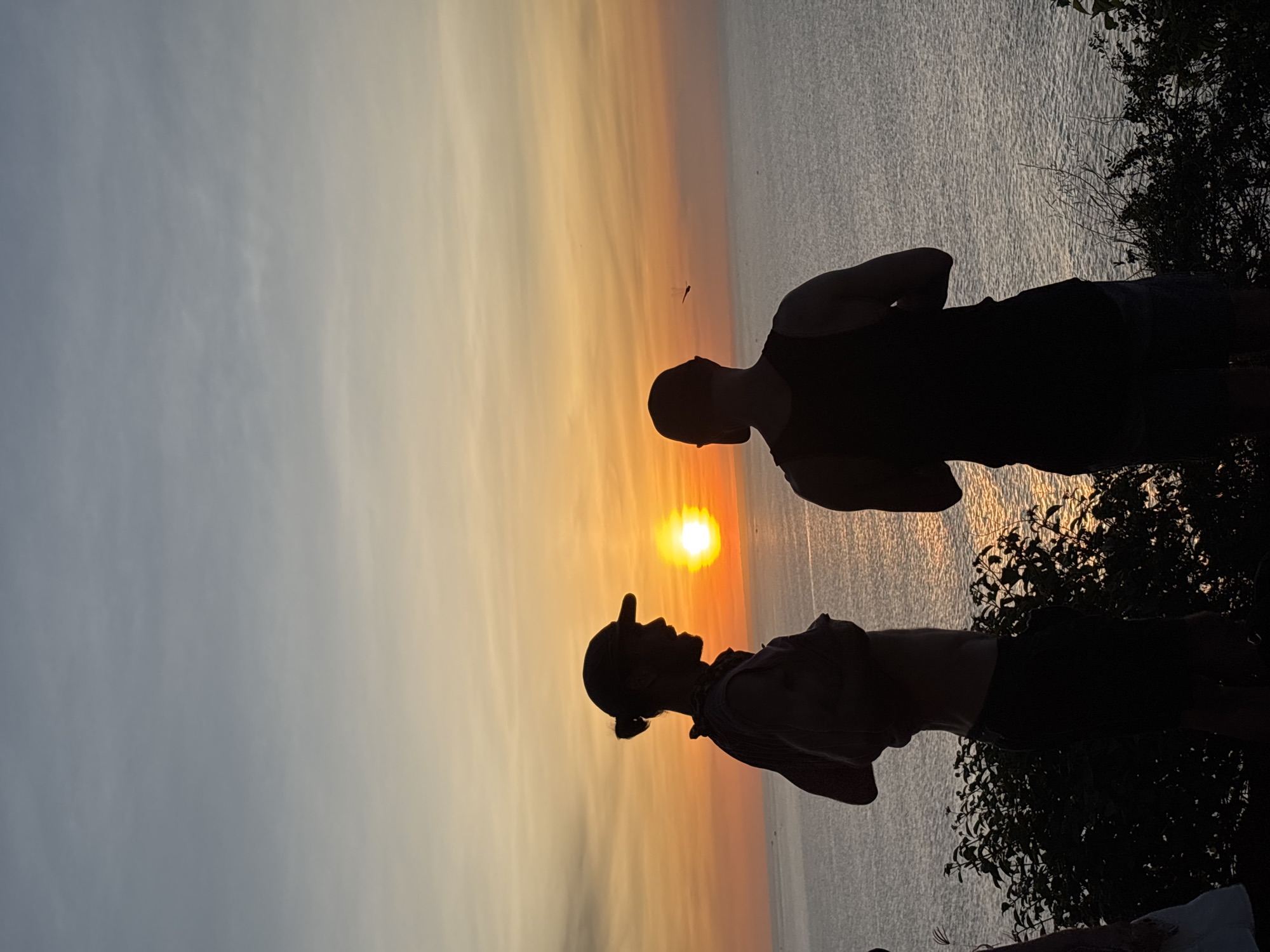 Two silhouetted men stand facing each other against a sunset over the ocean. One man wears a hat and has a ponytail; the other man wears a tank top. Trees are visible on the right. The location is Kuta Selatan, Indonesia, based on the likely location of a sunset over the ocean.