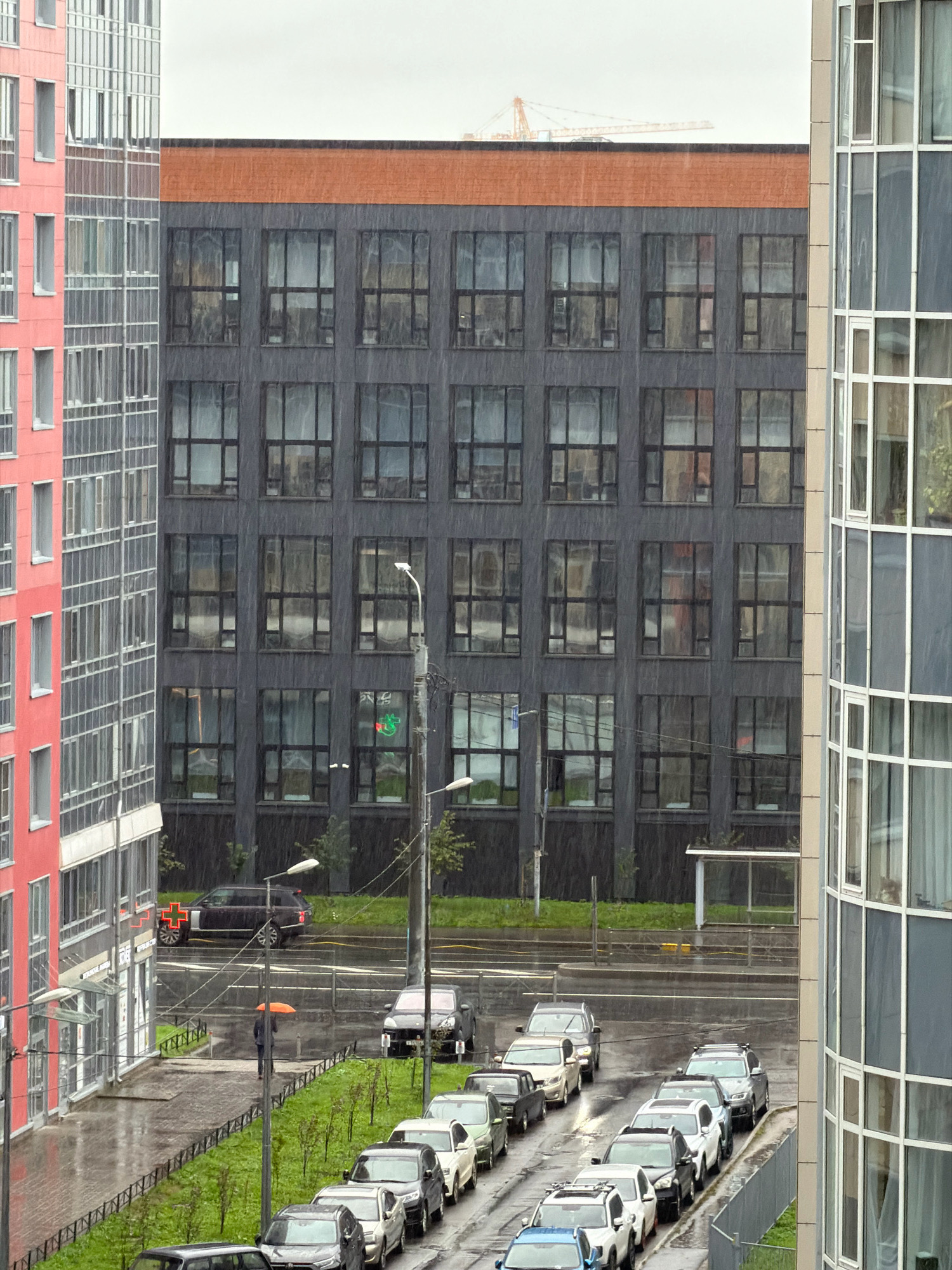 This is a medium-high angle exterior shot of a residential street in Saint Petersburg, Russia, during a rain shower. Multiple modern buildings are visible, including a tall pink building on the left, a dark gray multi-story building with numerous windows in the center, and a curved glass-fronted building on the right. The sky is overcast and the ground and buildings are wet, indicating rain. A line of cars is parked on the street in the foreground, with more vehicles visible further down the road. One person is walking with an orange umbrella. A red cross sign is visible on the pink building. A construction crane is visible in the distance behind the buildings.