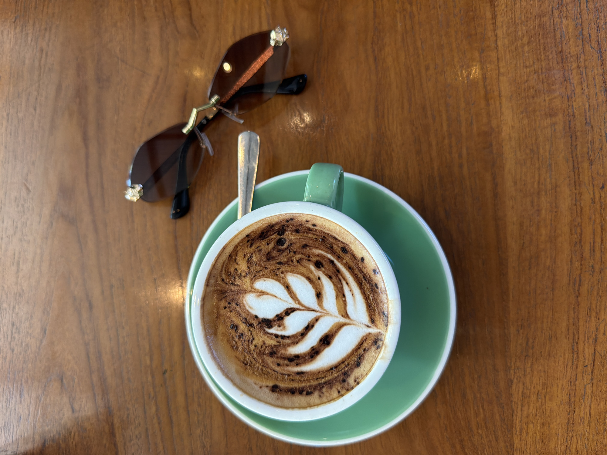 The image presents a high-angle view of a coffee cup, spectacles, and a spoon situated on a wooden surface. The coffee cup is green with a white interior, containing a latte with leaf-like latte art created in the foam. A small spoon is placed in the coffee. Adjacent to the coffee cup is a pair of brown-tinted rimless spectacles with ornamental golden flowers near the temples. The wooden surface has a warm tone and visible grain, possibly part of a table in a cafe or coffee shop. The location is Unknown, Unknown.