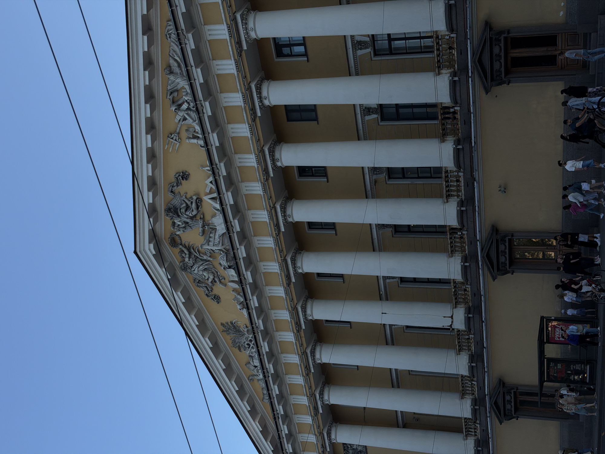 The image shows a wide, angled shot of the facade of a classical building. The building is painted in a light yellow color and features a prominent row of tall, white Corinthian columns. Above the columns, there is an ornate frieze decorated with mythological figures and tridents. The building has multiple windows with dark frames, and balconies with decorative railings. In the foreground, a street scene unfolds with people walking and standing near a bus stop shelter. A man on a bicycle is also visible. The sky is clear and blue. The overall impression is that of a grand, historic urban setting.