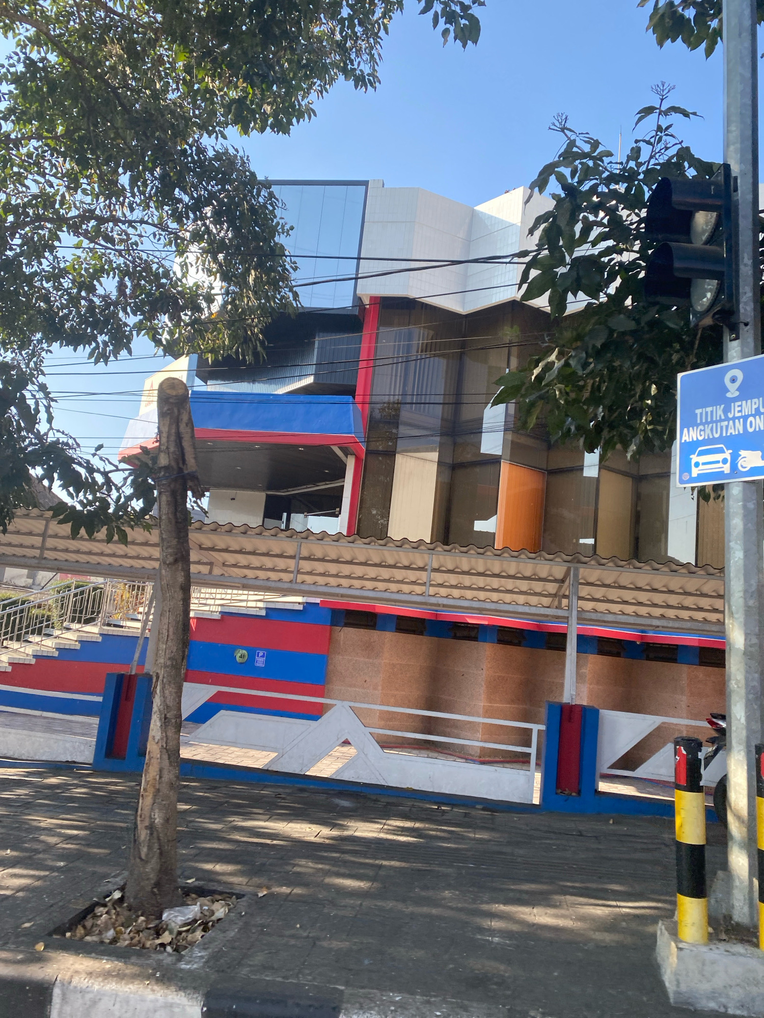 The image shows a building with a modern facade, featuring large windows and white and blue architectural elements. In the foreground, there is a sidewalk with a tree growing from a small patch of soil. A metal fence with red and blue accents runs along the property line, and a pedestrian walkway with stairs is visible behind it. A traffic light is mounted on a pole, and a blue sign indicates a pickup point for public transportation, showing icons for a car and a motorcycle. The sky is clear and blue. The scene appears to be in an urban environment, possibly a commercial area or an office complex.