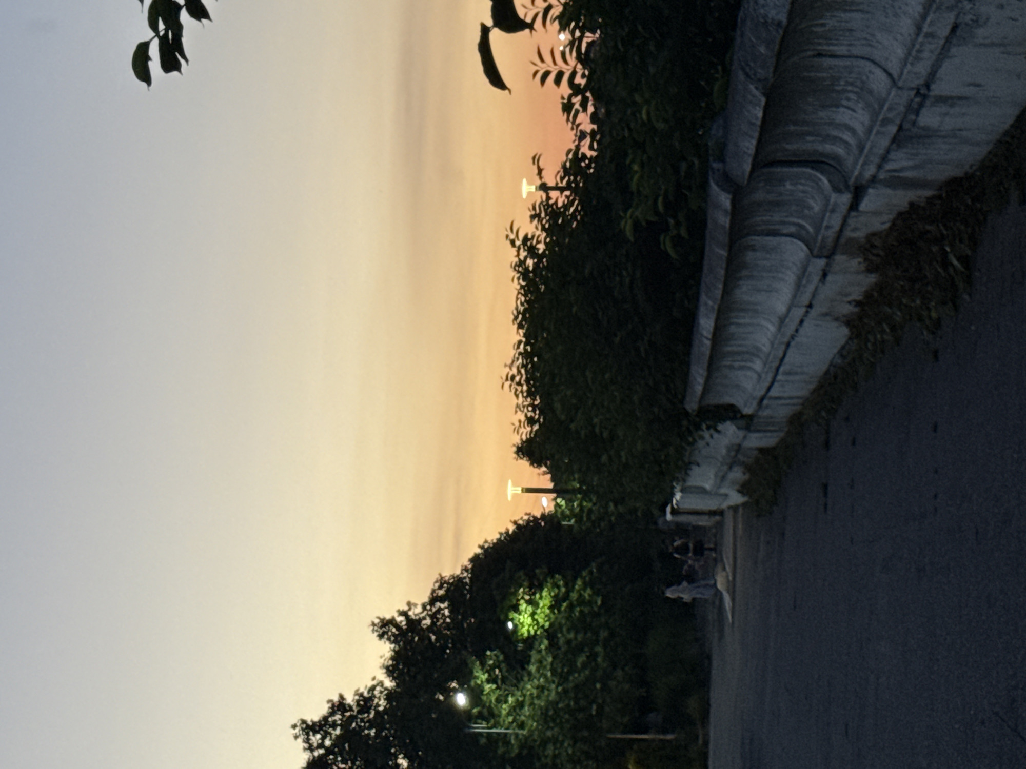 The image captures a serene evening scene in Kul'zeb, Russia, likely during sunset. The sky transitions from a pale grey at the top to a warm, peachy orange near the horizon, indicating the time of day. Silhouetted against this colorful sky are the dark leaves of a tree in the foreground and the lush foliage of trees lining a pathway.
The pathway itself appears to be paved and runs alongside a low concrete barrier. Beyond the barrier, a dense line of trees creates a natural boundary. Several lampposts are visible along the pathway, some emitting a soft glow, suggesting the onset of twilight. In the distance, a few individuals are walking or standing on the pathway, their forms indistinct in the low light, adding a sense of human presence to the tranquil setting. The overall mood is peaceful and contemplative, with the natural beauty of the sunset complemented by the subtle illumination of the pathway lights. No visible text is present in the image.