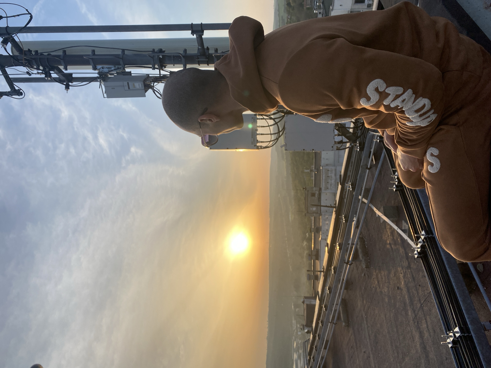 A person wearing sunglasses and a brown "Stadia" hoodie is on top of a high structure, possibly a communications tower. The sun is low on the horizon, creating a hazy, golden light. The tower is made of metal with various cables and boxes attached. The city of Pleven, Bulgaria is visible in the background.