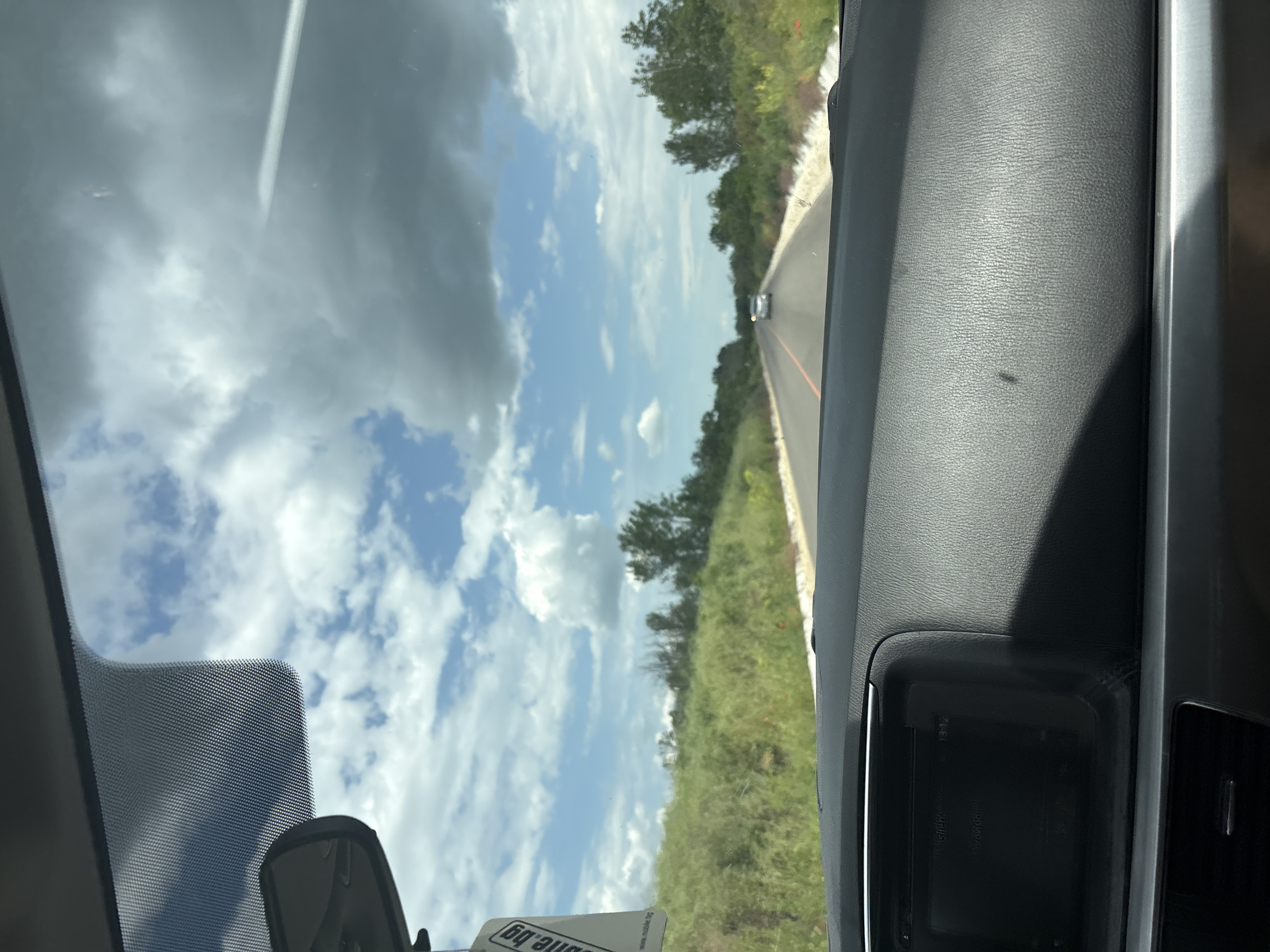 The view from inside a vehicle traveling on a paved road is seen. The sky is visible through the windshield with clouds. Trees and vegetation line the roadside. Another vehicle is traveling ahead on the same road. The dashboard is visible in the foreground. There is no indication this image is from Aglen, Bulgaria.