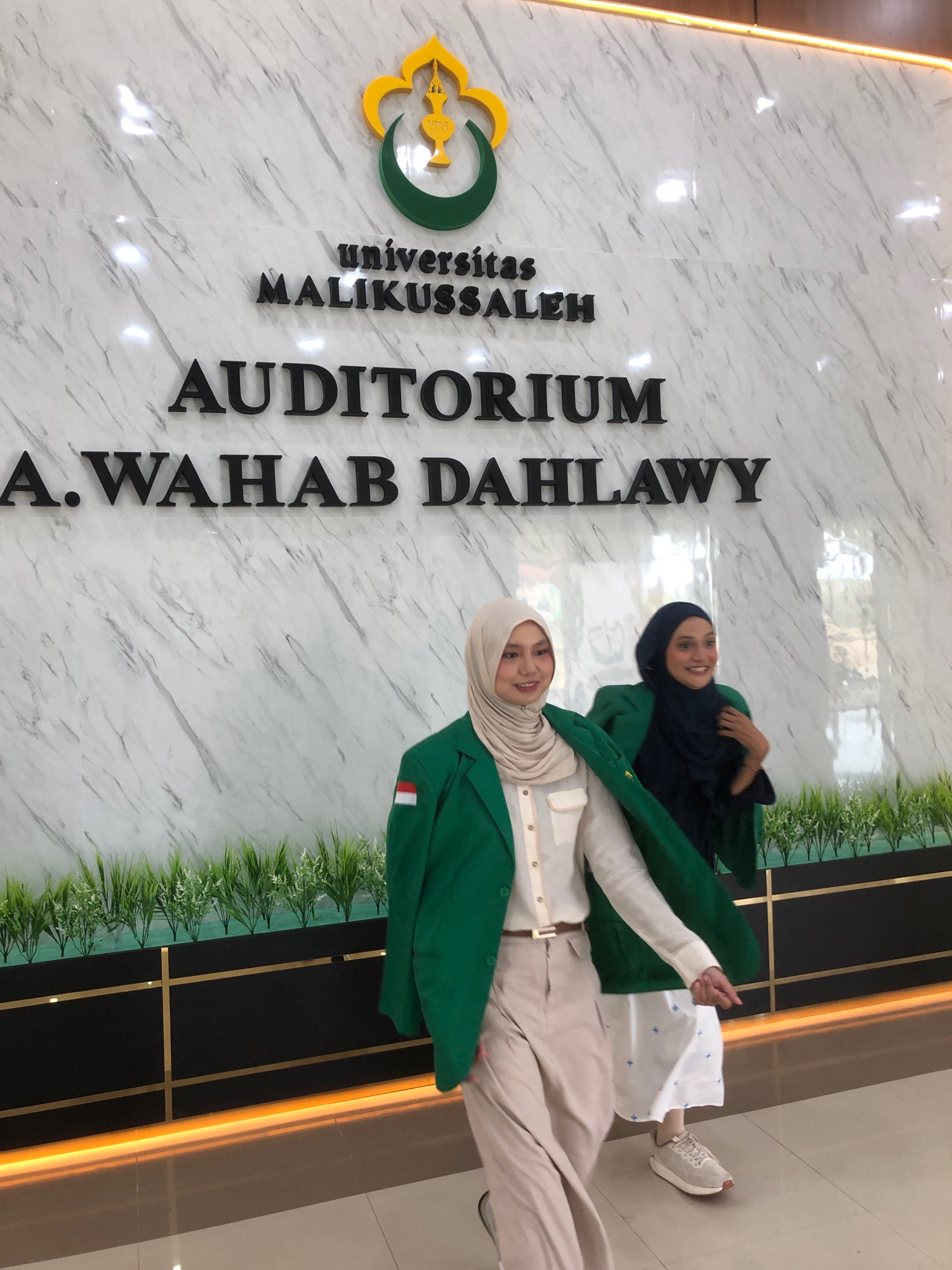Two women are posing in front of a marble wall with text that reads "Universitas Malikussaleh Auditorium A. Wahab Dahly" in Reuleut Timu, Indonesia. The woman in the foreground is wearing a light-colored hijab, a white shirt, beige pants, and a green blazer with an Indonesian flag patch on the sleeve. The woman behind her is wearing a dark blue hijab and a dark green blazer over a white dress. Both women are smiling and looking towards the camera. The environment includes decorative plants in planters and a warm-toned light strip along the base of the wall.