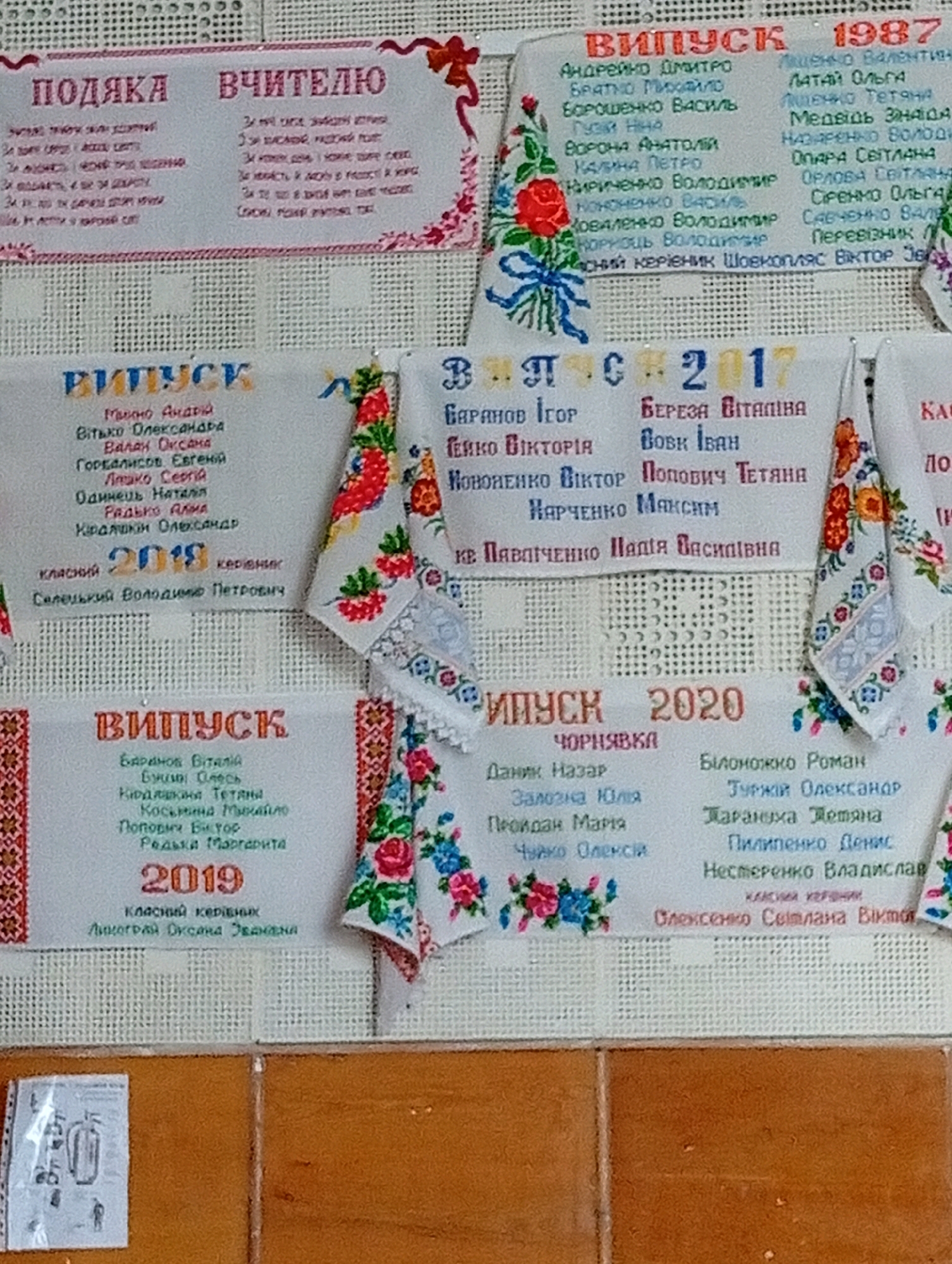 The image displays a collection of embroidered banners, likely for graduation or a celebratory event, arranged on a textured background. The banners are adorned with Ukrainian text and decorative floral patterns. One banner at the top left has the heading "ПОДЯКА ВЧИТЕЛЮ" (Gratitude to the Teacher). Several other banners are marked "ВИПУСК" (Graduation) followed by years such as 1987, 2017, 2018, 2019, and 2020. Each graduation banner lists names, presumably of graduates and possibly class teachers. The banners are hung on what appears to be a wall, with a glimpse of a wooden surface at the bottom of the image. The overall scene suggests a commemorative display in a school or community setting. No specific geographical location within "Unknown, Unknown" can be determined from the visual information.