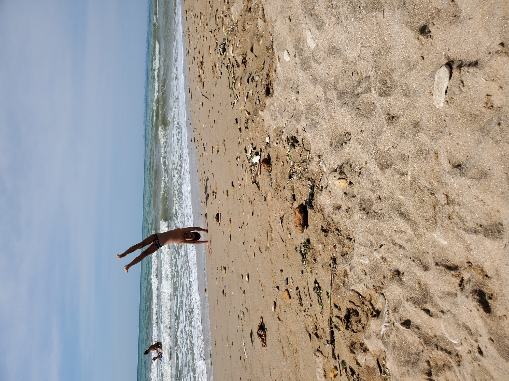 A young person is performing a handstand on a sandy beach with the sea in the background. The person is wearing dark swim trunks. There are waves gently rolling onto the shore. In the distance, two other people are visible in the shallow water. The beach is littered with small stones and some dried seaweed. The sky is partly cloudy, and a distant city skyline can be seen on the horizon.