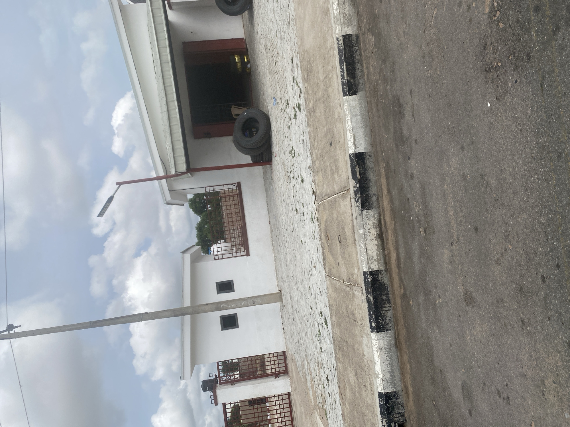 A concrete roadside with black and white painted curb runs along the bottom of the frame. Behind the curb is a paved area with interlocking gray concrete pavers, some of which are cracked and have weeds growing through them. In the middle ground, there is a white building with a large open doorway. In front of the building, a stack of three black tires is visible. The building appears to be a commercial establishment, possibly a tire shop. To the left of the building, there is a smaller white structure with two dark windows, resembling a guard post or small office. A red metal gate with a diamond pattern is open to the left of this structure, and a red metal fence runs along the front of the property. A utility pole with overhead wires is present in the left midground. The sky above is filled with white, puffy clouds against a pale blue background. The overall scene depicts a commercial area on a street in Jalingo, Nigeria.