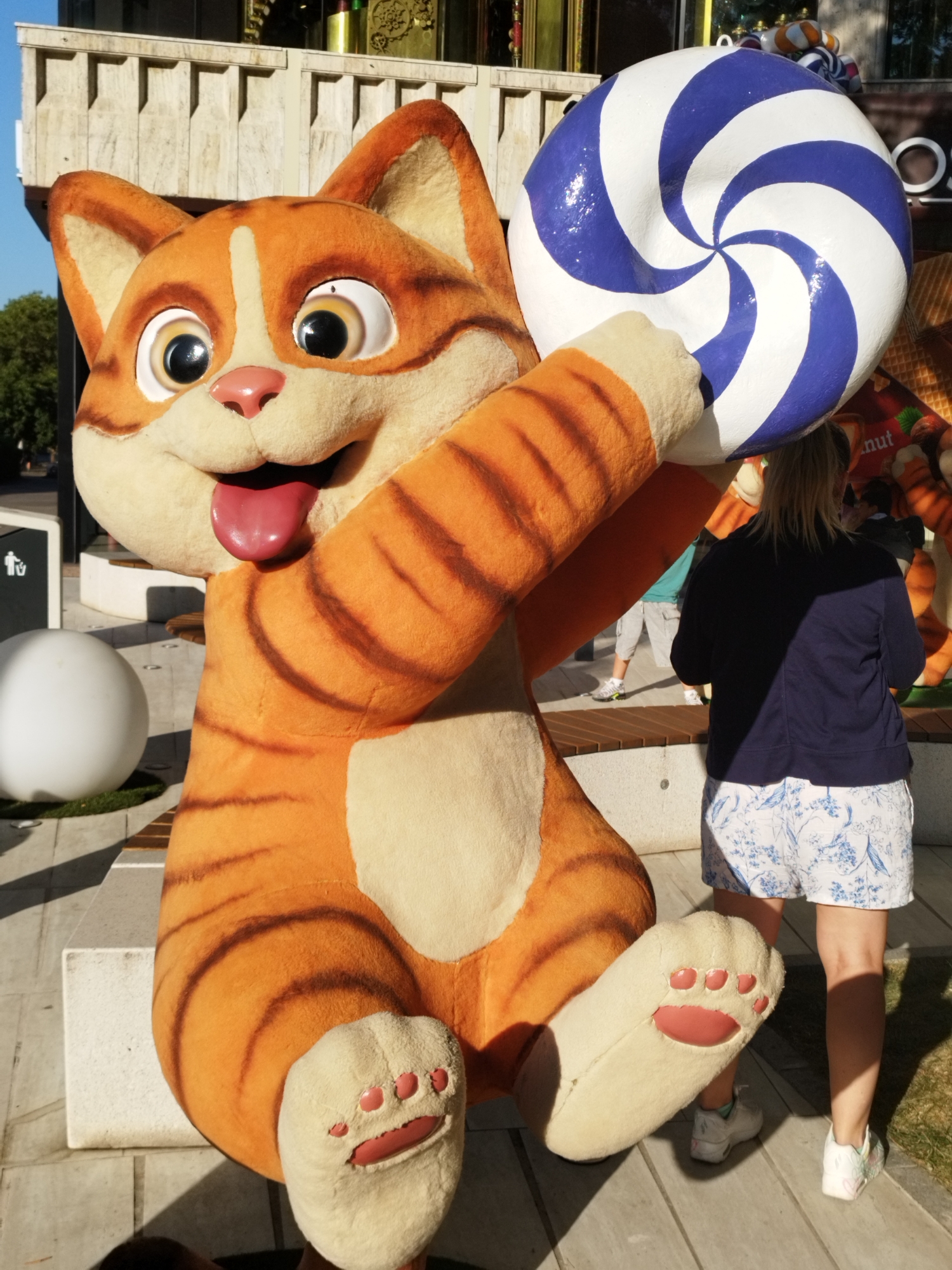 This is a photograph taken outdoors, possibly in a public space like a park or an entertainment district, in Bucharest, Romania. The main subject is a large, plush statue of an orange and white striped cat character. The cat is depicted in a playful pose, holding a giant blue and white swirled lollipop in its right paw. Its mouth is open, with its pink tongue sticking out.
In the background, a woman with blonde hair is partially visible from the back, wearing a dark blue jacket and white shorts with a blue floral pattern. She is standing to the right of the cat statue. Further in the background, other people and elements of the urban environment are visible, including a building with decorative architectural features, a white spherical object, benches, and trees. The lighting suggests it is daytime, with sunlight casting shadows. The overall impression is of a lively and possibly whimsical setting.