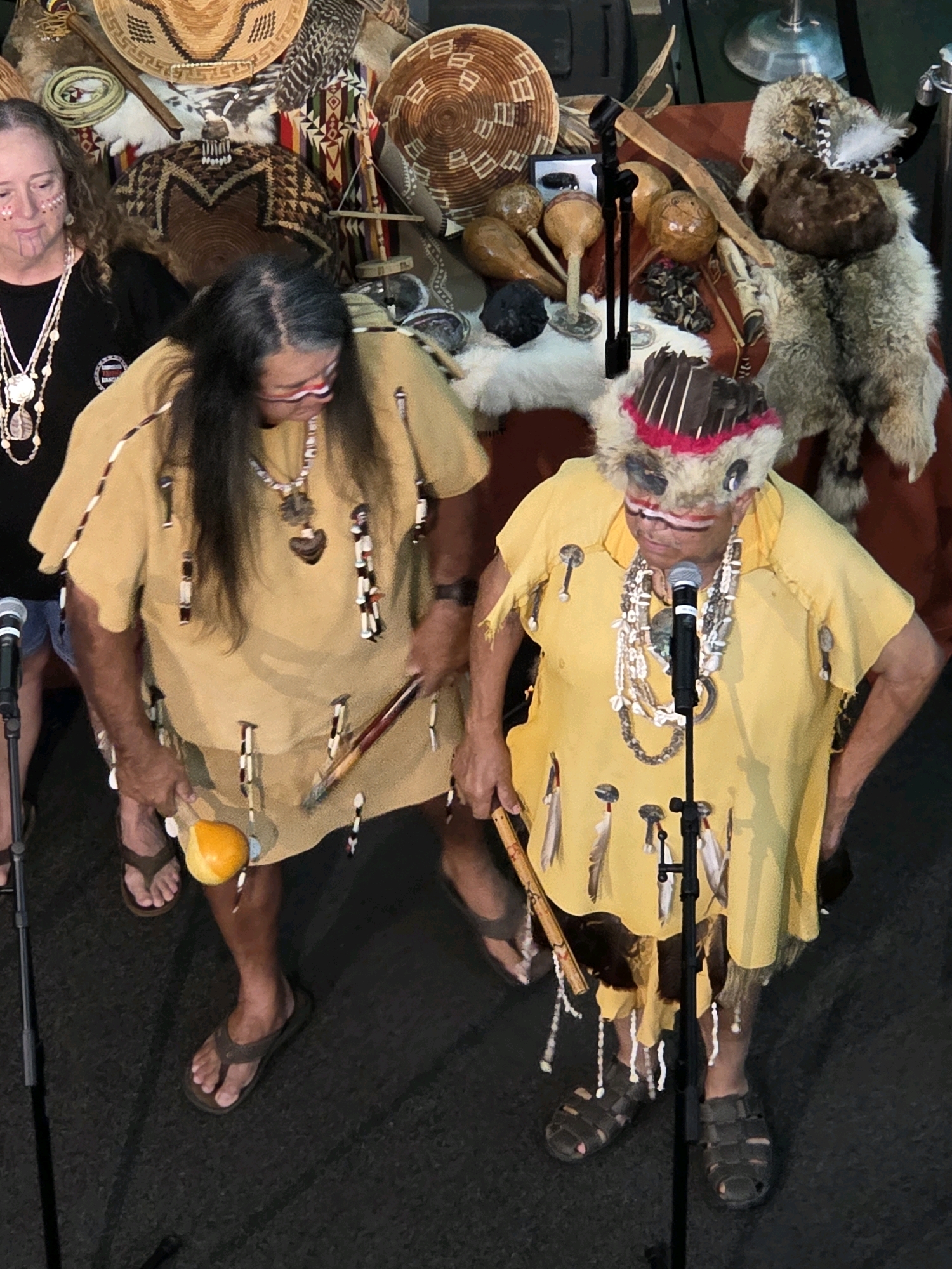 Two individuals, appearing to be of Native American heritage, are standing and performing in front of a display of cultural artifacts. The individual on the left wears a black t-shirt and a tan ceremonial robe adorned with beads and feathers. They are holding a yellow gourd rattle and a wooden implement. The individual on the right also wears a tan ceremonial robe, decorated similarly, and a headdress with feathers and fur. They are holding a wooden implement and are standing near a microphone. The backdrop consists of various woven baskets, furs, and other traditional items. The scene appears to be an indoor cultural event, possibly at the Long Beach Museum of Art in Long Beach, California.