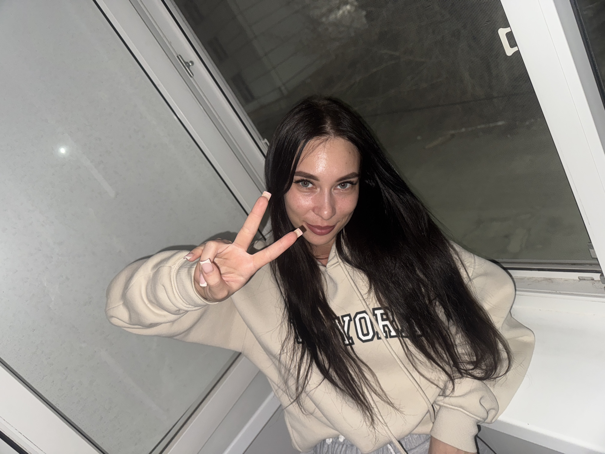 A young woman is posing indoors. She has long, dark hair and is wearing a light-colored hoodie with the word "YORK" printed on it. She is making a peace sign with her right hand. The background appears to be a window with a screen and a view to the outdoors at night. Based on other available information, the location is Chaikovsky, Russia.