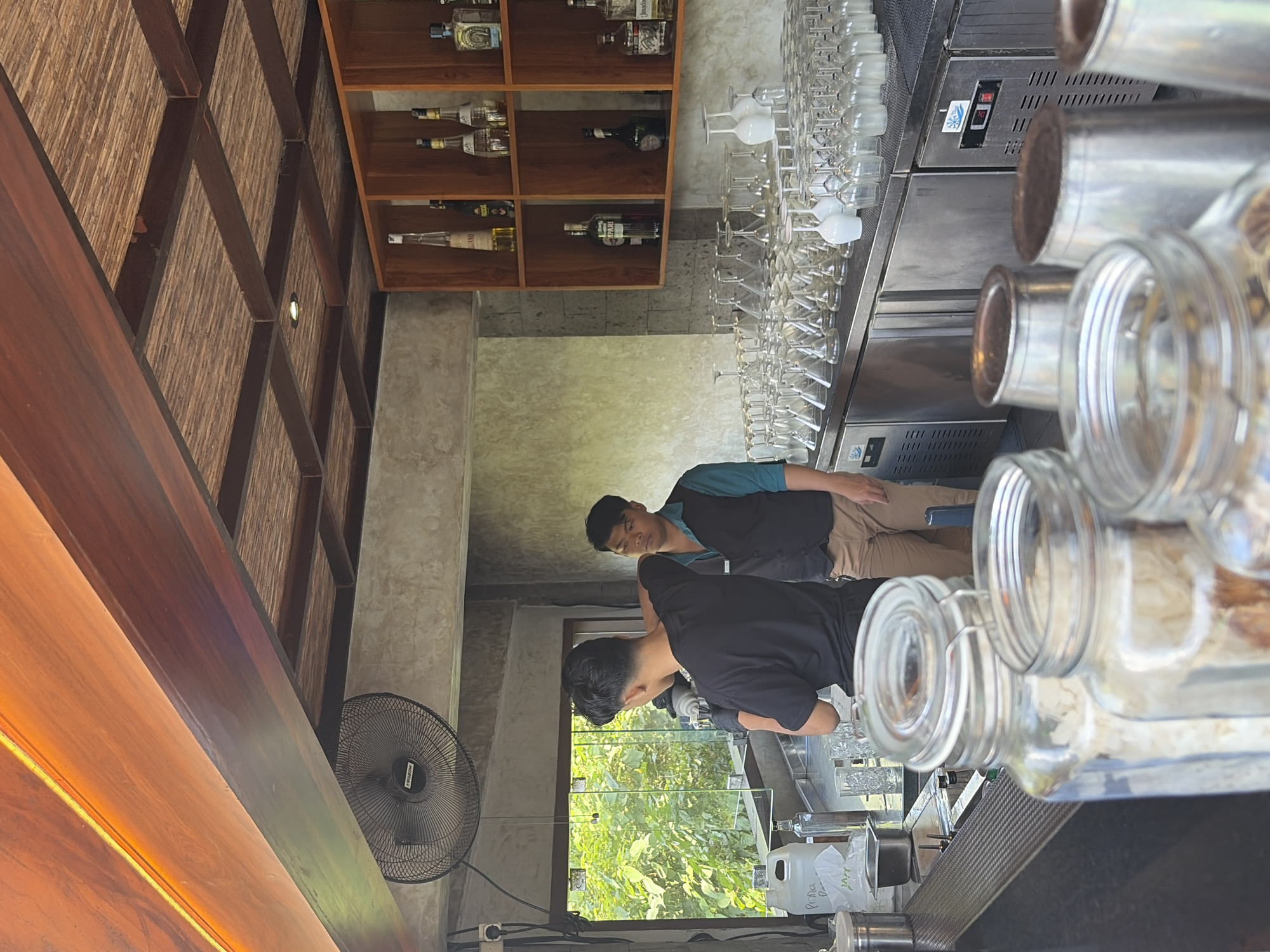 The image depicts an indoor bar area with two men, presumably bartenders, working behind a counter. The man on the left, wearing a black t-shirt and black gloves, is focused on preparing something at the bar. The man on the right, in a blue shirt and black vest, stands by observing. The bar is equipped with numerous glassware, including wine glasses and martini glasses, neatly arranged on shelves and the counter. Bottles of liquor are visible on shelves in the background. The overall ambiance suggests a tropical setting, possibly a restaurant or cafe, with natural wood accents on the ceiling and an open window revealing lush green foliage outside. The presence of a fan mounted on the ceiling further contributes to the tropical feel. The scene is set in Tegallalang, Indonesia.