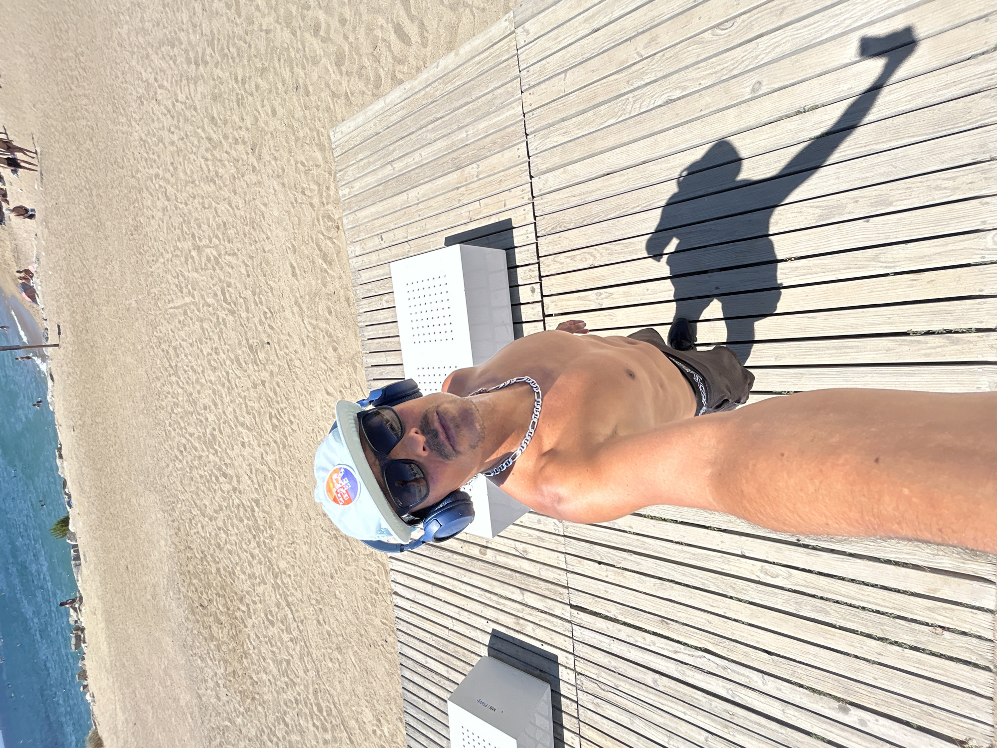 A man wearing a light blue baseball cap with an emblem, dark sunglasses, and blue headphones takes a selfie on a wooden boardwalk at a beach in Barcelona, Spain. He is shirtless and wearing dark shorts. The beach is sandy with the Mediterranean Sea in the background. There are several white cubic structures on the boardwalk. The sun is casting a long shadow of the man on the boardwalk. In the distance, people can be seen on the beach and in the water.