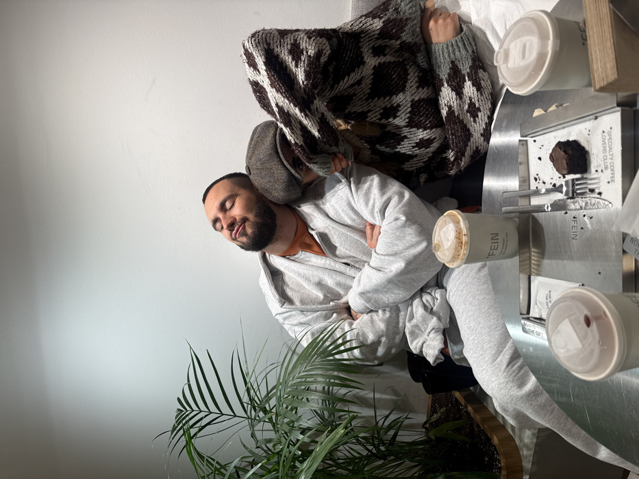 A man with a beard and closed eyes leans against a woman wearing a brown and white sweater. She has a hat on. He's wearing a gray sweatsuit. A plant sits below them. They are sitting at a table with coffee cups and a piece of cake. The table has cutlery and several cards on it.