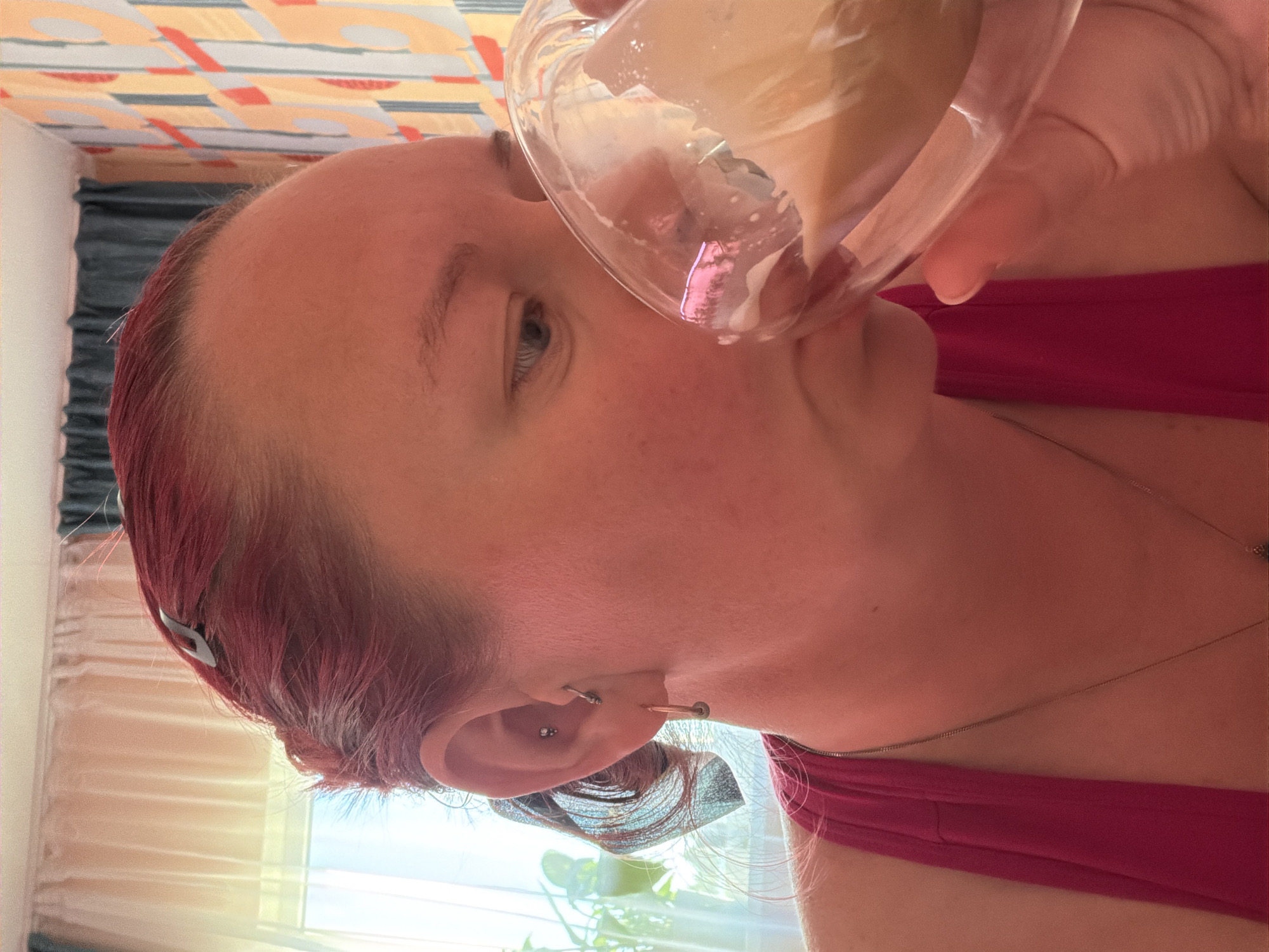 A person is shown in a close-up shot, holding a glass to their lips, presumably drinking. The person has light skin, light eyes, and reddish-purple hair with a hair clip. They are wearing a pink top and a delicate necklace. The background includes a window with light-colored curtains, and what appears to be a patterned ceiling or wall. There is no information to determine the location is in the city of Chaikovsky, Russia.