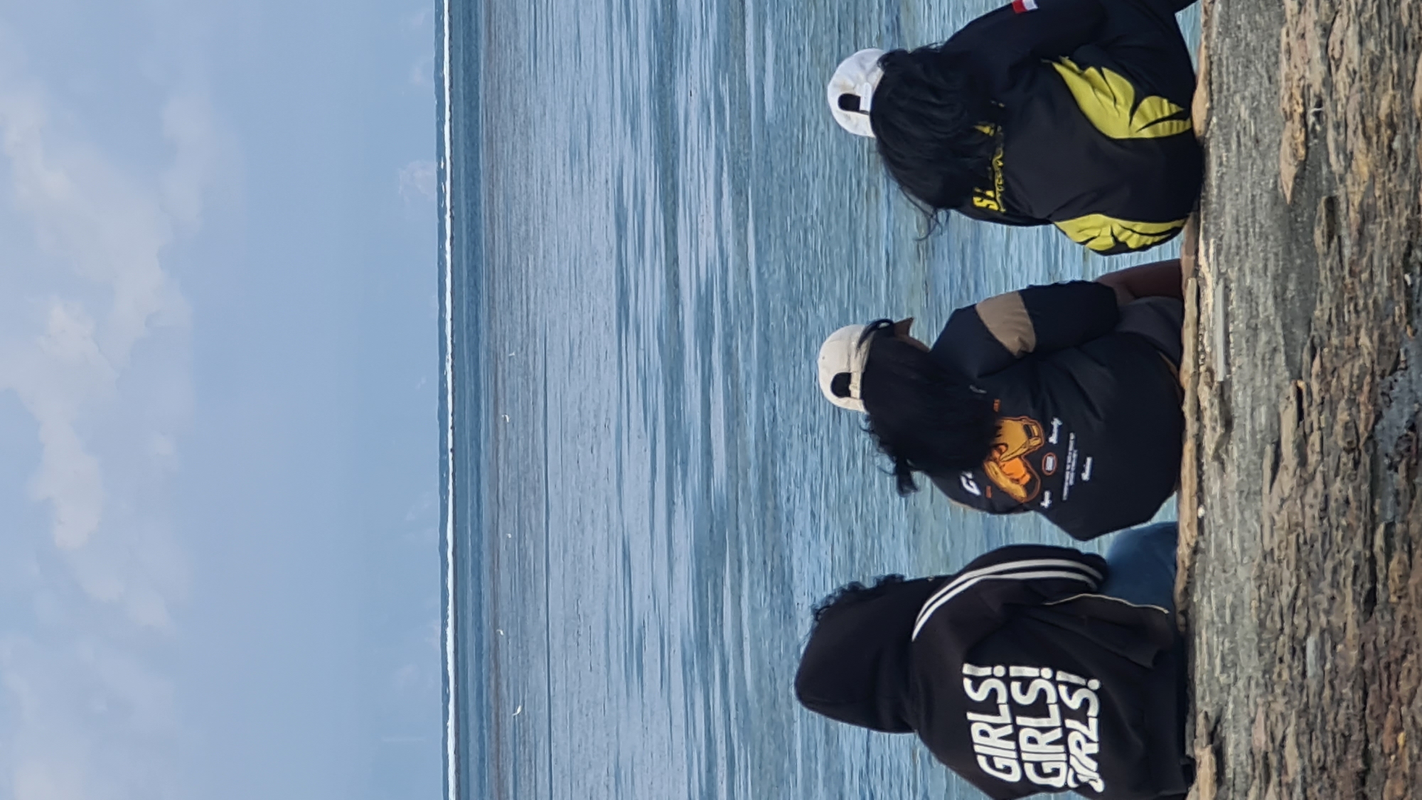 Three individuals are seated on a rocky outcrop overlooking a calm body of water. The individual on the left wears a black hoodie with white stripes on the sleeves and text on the back that reads "GIRLS! GIRLS! GRLS!". The middle individual wears a black jacket with a brown panel on the sleeve and a graphic design on the back featuring an orange baseball glove. The individual on the right wears a black jacket with yellow wing-like designs on the sleeves and a red and white flag patch on the right sleeve. The water is a calm blue with a faint horizon line visible in the distance. The sky is partly cloudy, with shades of blue and white. This scene is consistent with a view from a coastal area in Denpasar, Indonesia, likely near a beach or seaside promenade.