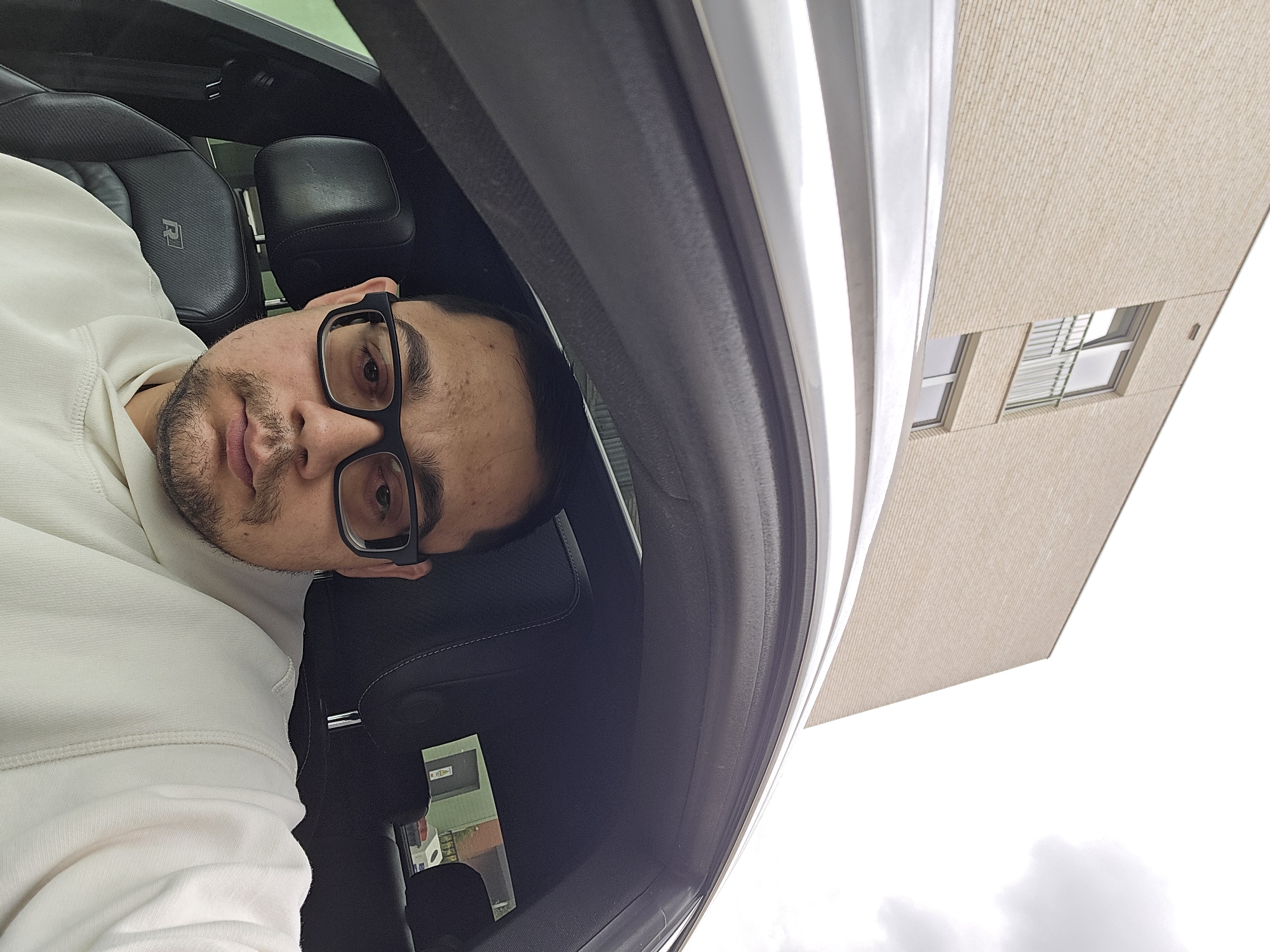 A medium shot captures a man with glasses and a goatee sitting in the driver's seat of a car, facing the camera. The car's interior is visible, including a black leather seat with an "R" logo and stitching. Outside the car, a modern building constructed with light-colored brickwork is seen, featuring several windows and a balcony. The sky above is overcast with visible clouds. The scene is likely in Ghent, Belgium, suggested by the architectural style. The man is wearing a light-colored hoodie.