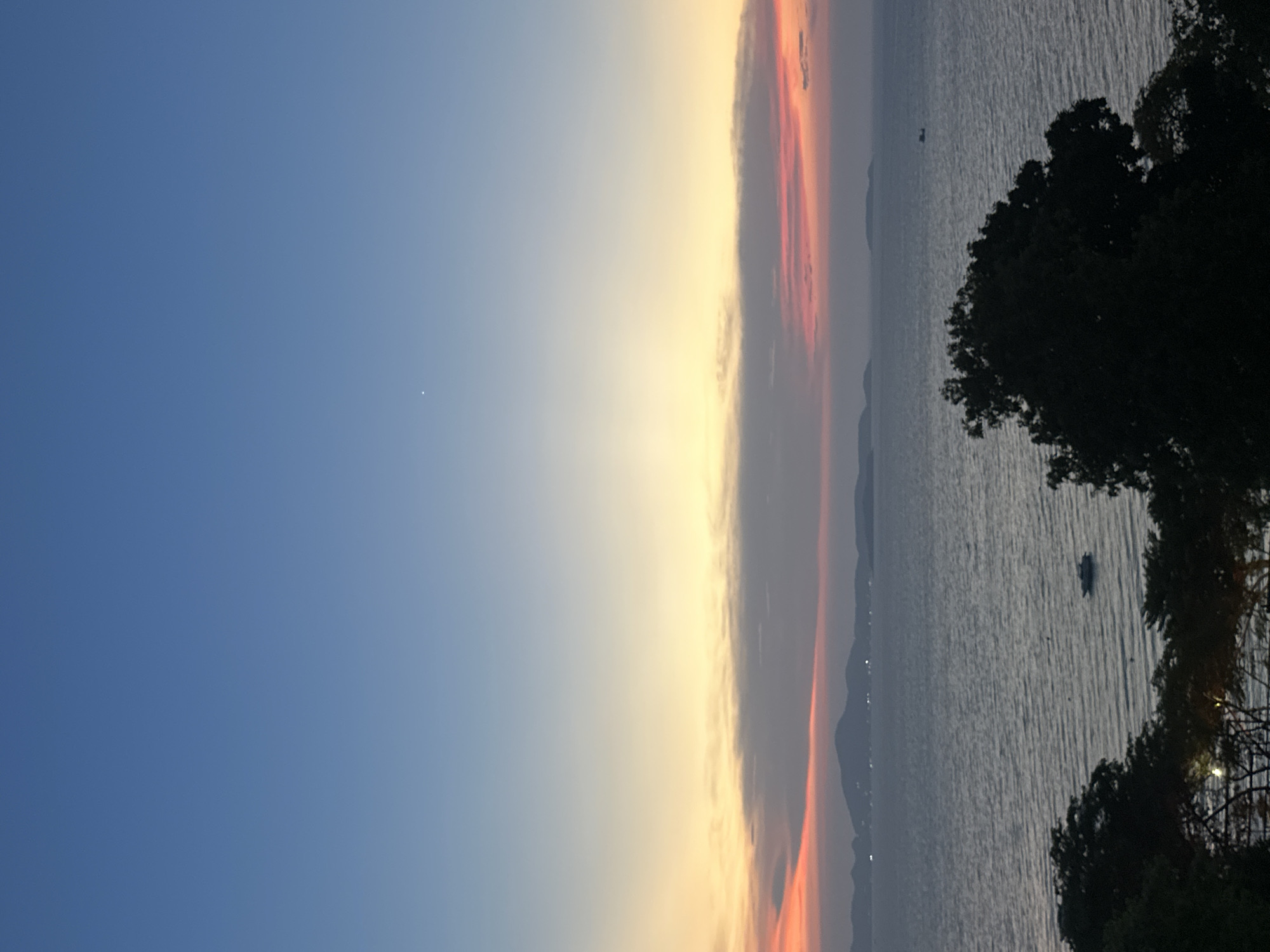 The image shows a sunset over the Gulf of Thailand, likely taken from a vantage point in or near Pattaya City, Thailand. The sky transitions from a clear blue on the left to a pale orange-yellow near the horizon, with streaks of red and grey clouds. Below the horizon is a body of water that is reflecting the sky and clouds. The silhouettes of hills or islands are faintly visible on the horizon of the water. On the right side of the image, there are blurred outlines of trees. There are a few small vessels in the water. A tiny bright dot might represent a celestial object such as a star or planet in the sky.