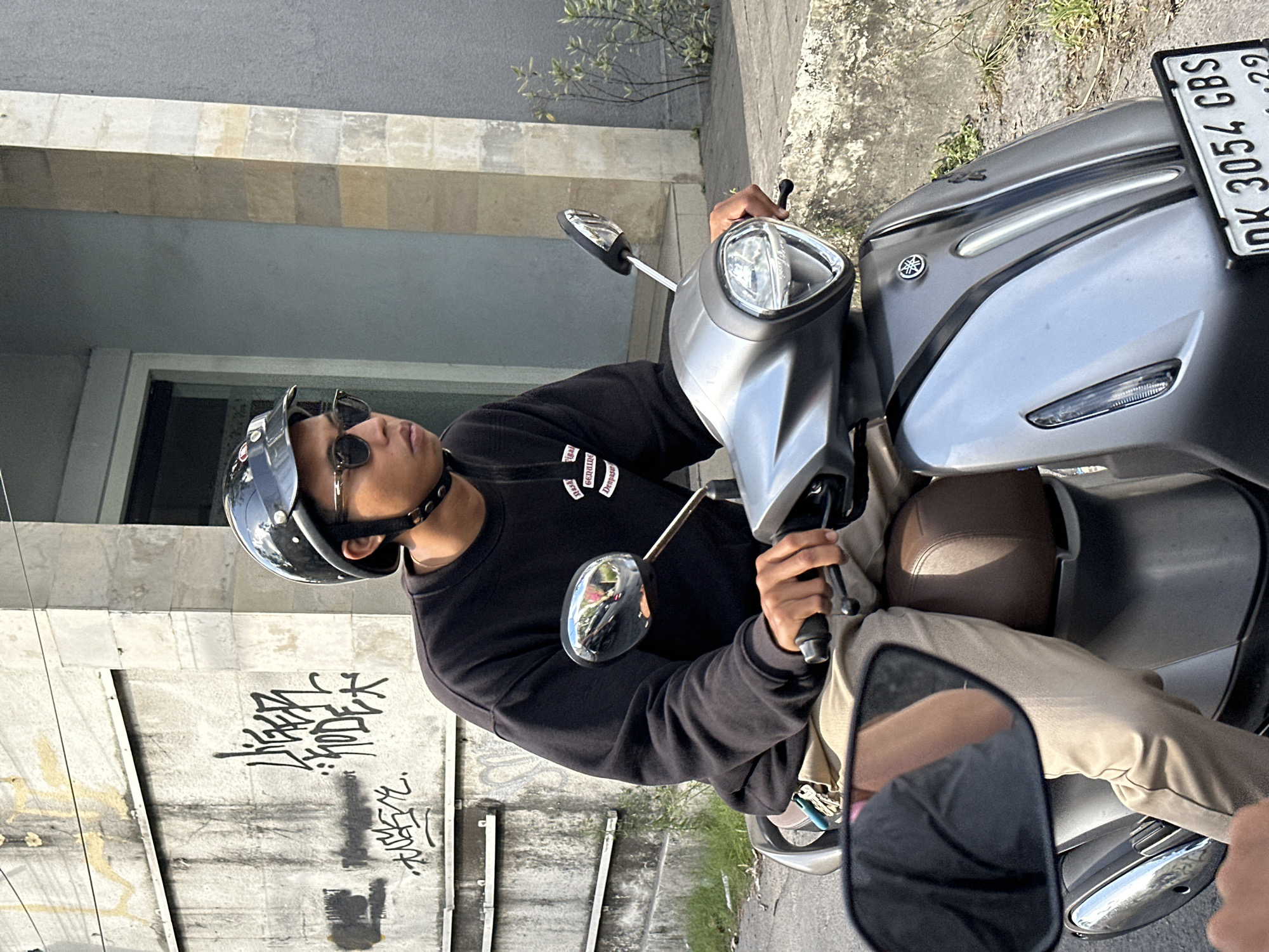A young man wearing a black helmet, sunglasses, and a black sweatshirt rides a silver Yamaha scooter. He is looking to his right. The scooter has a license plate that reads "DK 3054 CBS". The background includes a concrete wall with graffiti on it and a building. The scene appears to be on a street in North Kuta, Indonesia.